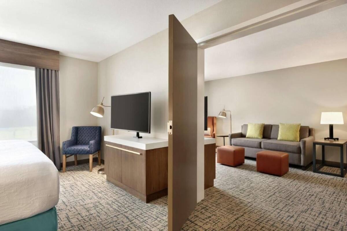 Ein Hotelzimmer mit Bett, Schreibtisch mit Flachbildfernseher, Stuhl, Sofa, zwei Hocker und einer Lampe.