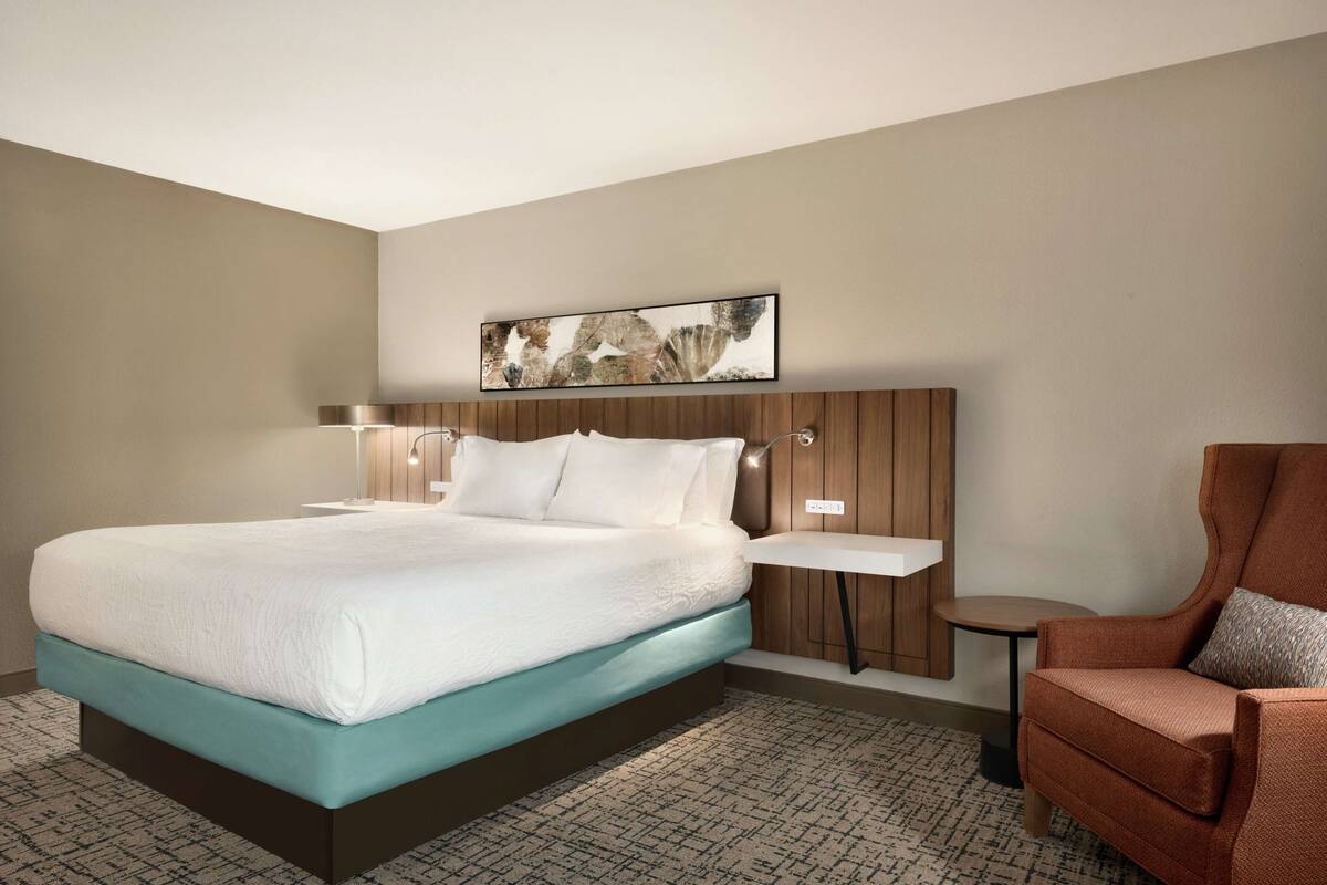 Ein Hotelzimmer mit einem großen Bett, einem kleinen Beistelltisch, einem Sessel und einem Wandbild.