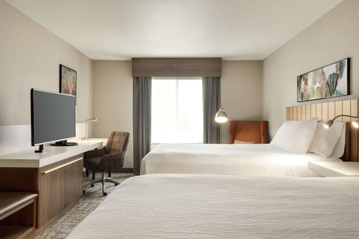 Ein Hotelzimmer mit zwei Betten, einem Schreibtisch, einem Flachbildfernseher, einem Sessel und einem großen Fenster mit Vorhängen.