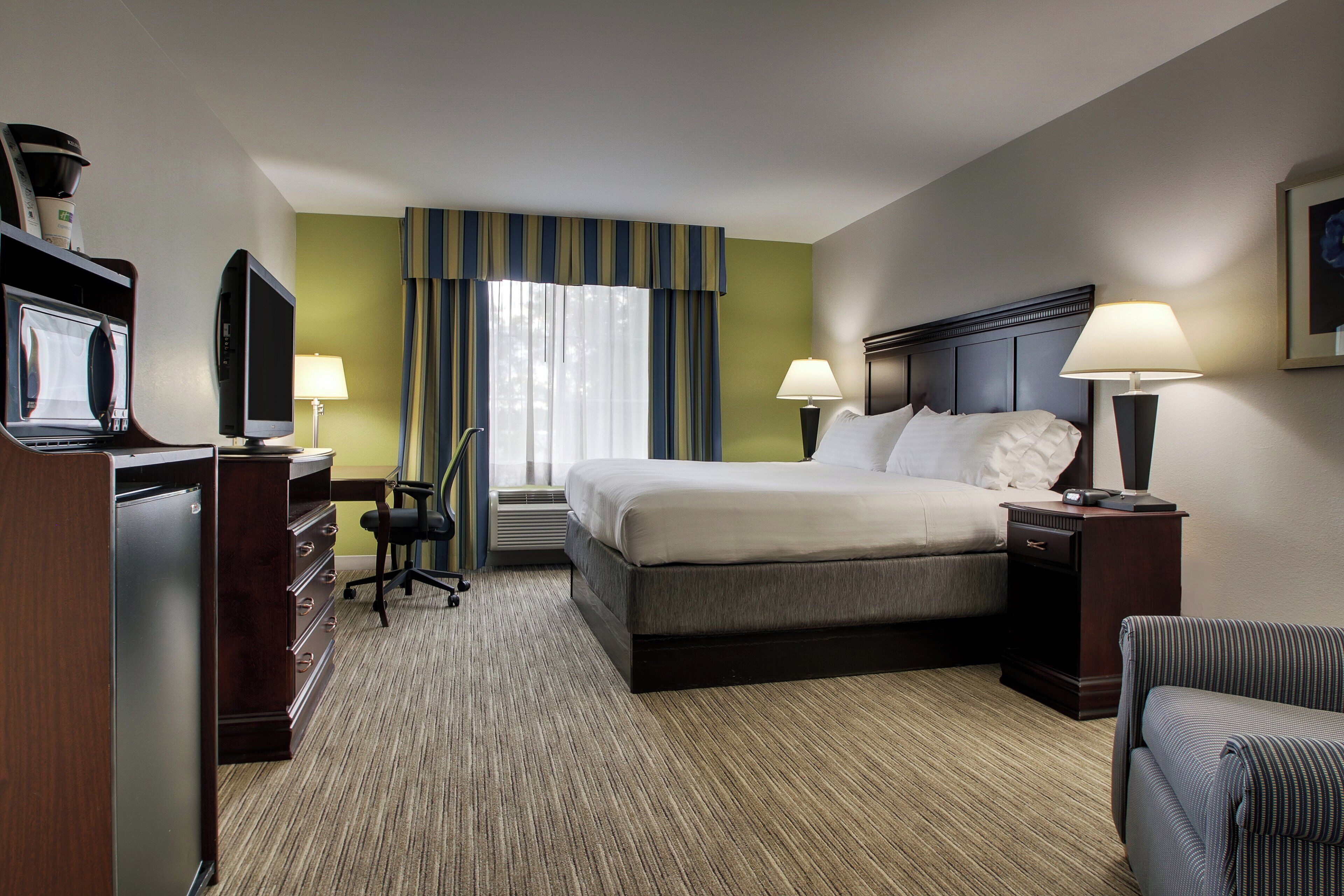 Quarto de hotel com cama, duas luminárias de cabeceira, escrivaninha com televisão, uma cadeira e janela com cortinas.
