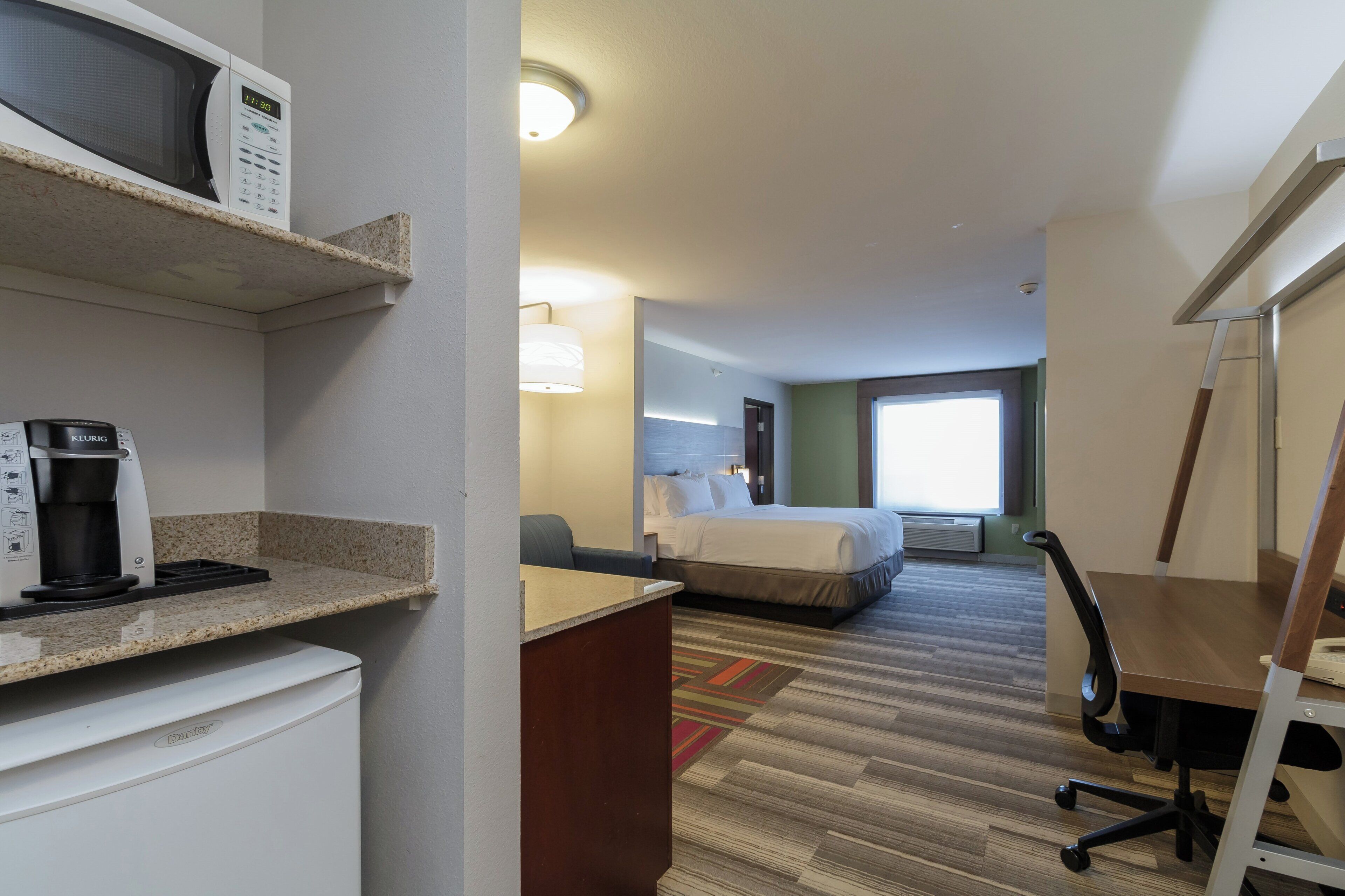 A hotel room with a bed, a desk with a coffee maker, a chair, and a lamp.
