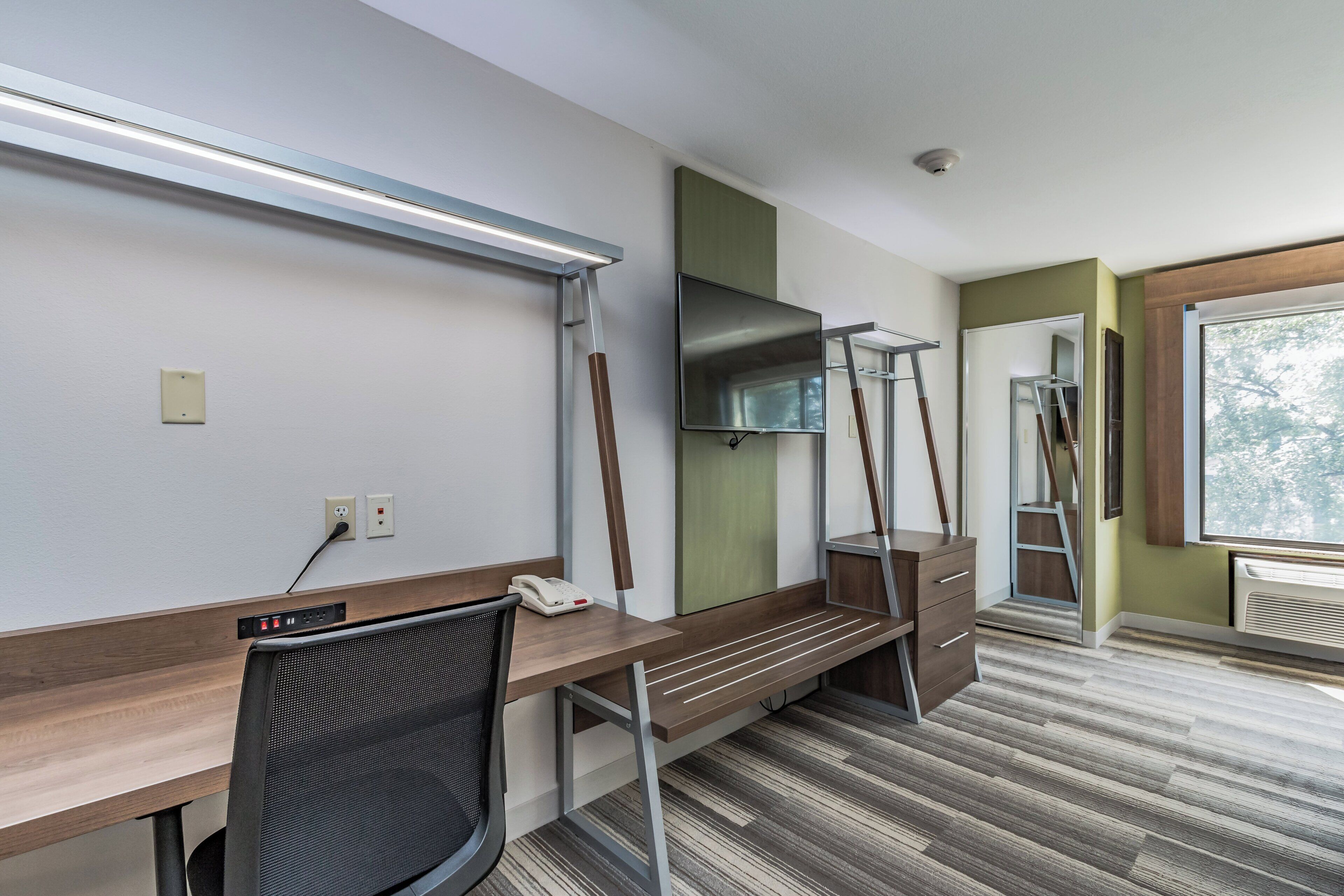 A hotel room with a desk, chair, flat-screen TV, telephone, and a window with a view of trees.
