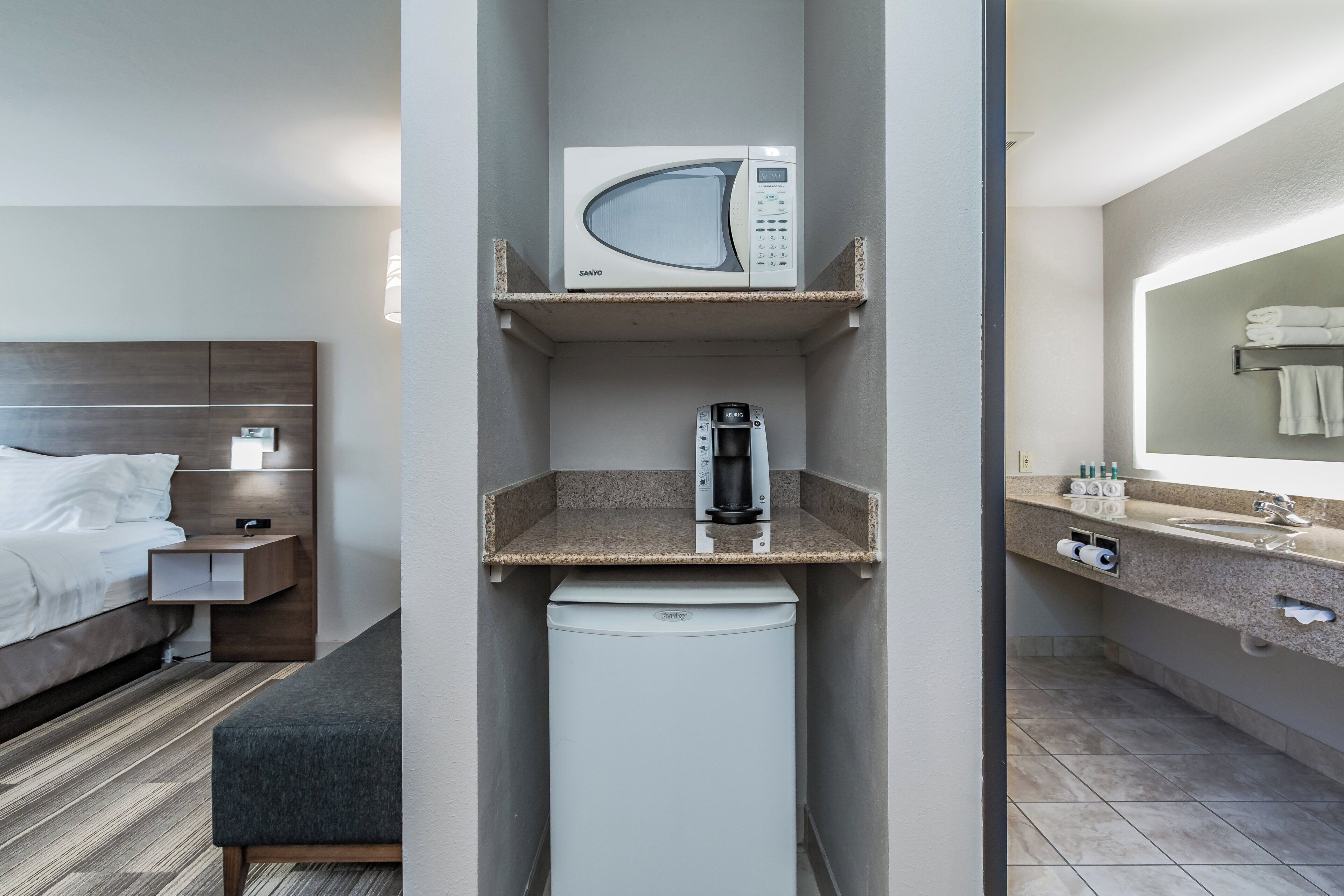 A hotel room with a bed, a nightstand, a microwave, a coffee maker, a small refrigerator, and a bathroom with a sink and mirror.