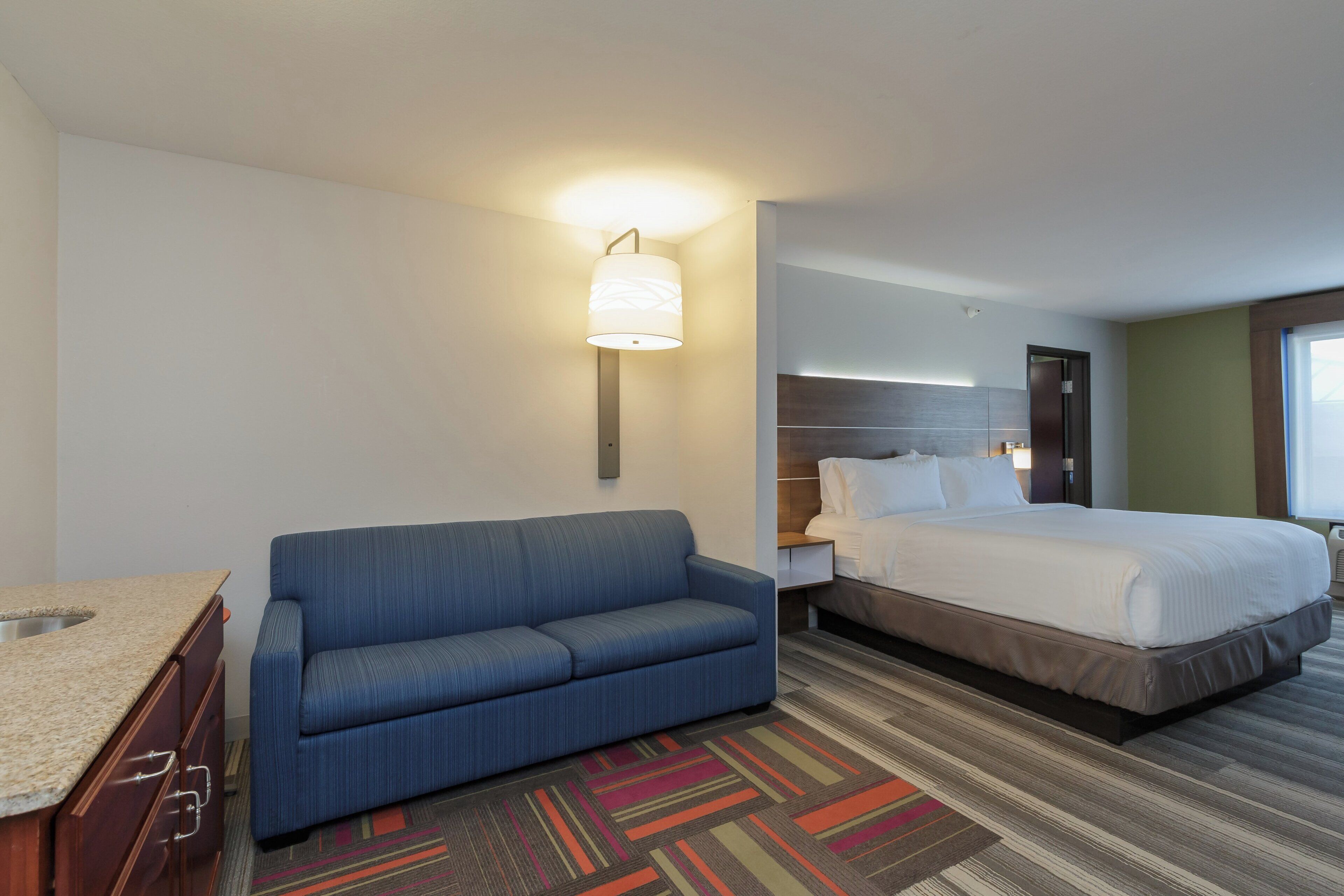 Una habitación de hotel con sofá, cama, mesita de noche y lámpara de pared.
