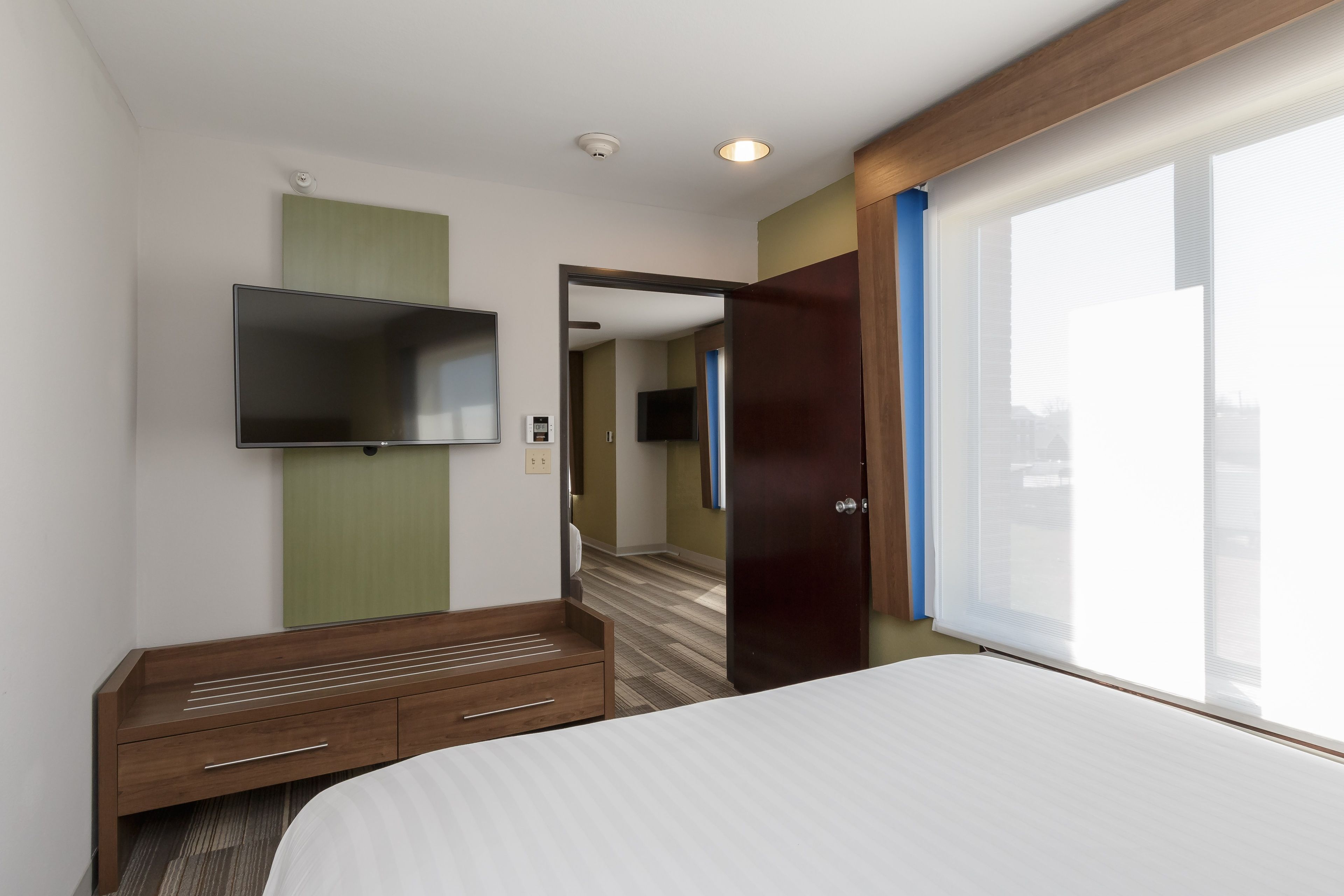 A hotel room with a bed, a TV mounted on the wall, a wooden dresser, and a door leading to another room.