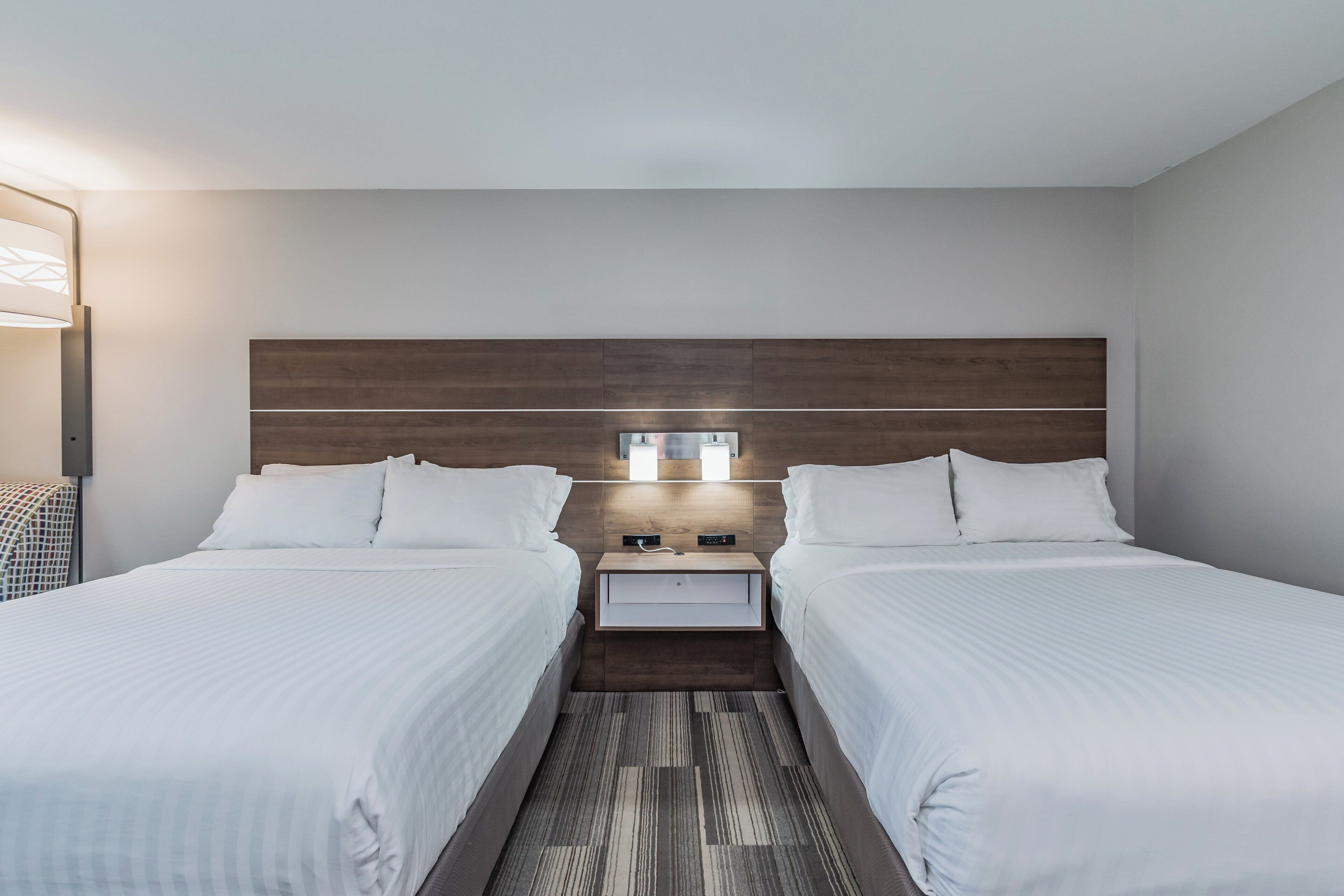 Une chambre d’hôtel avec deux lits, une tête de lit en bois, une table de chevet avec une lampe et un luminaire fixé au mur.