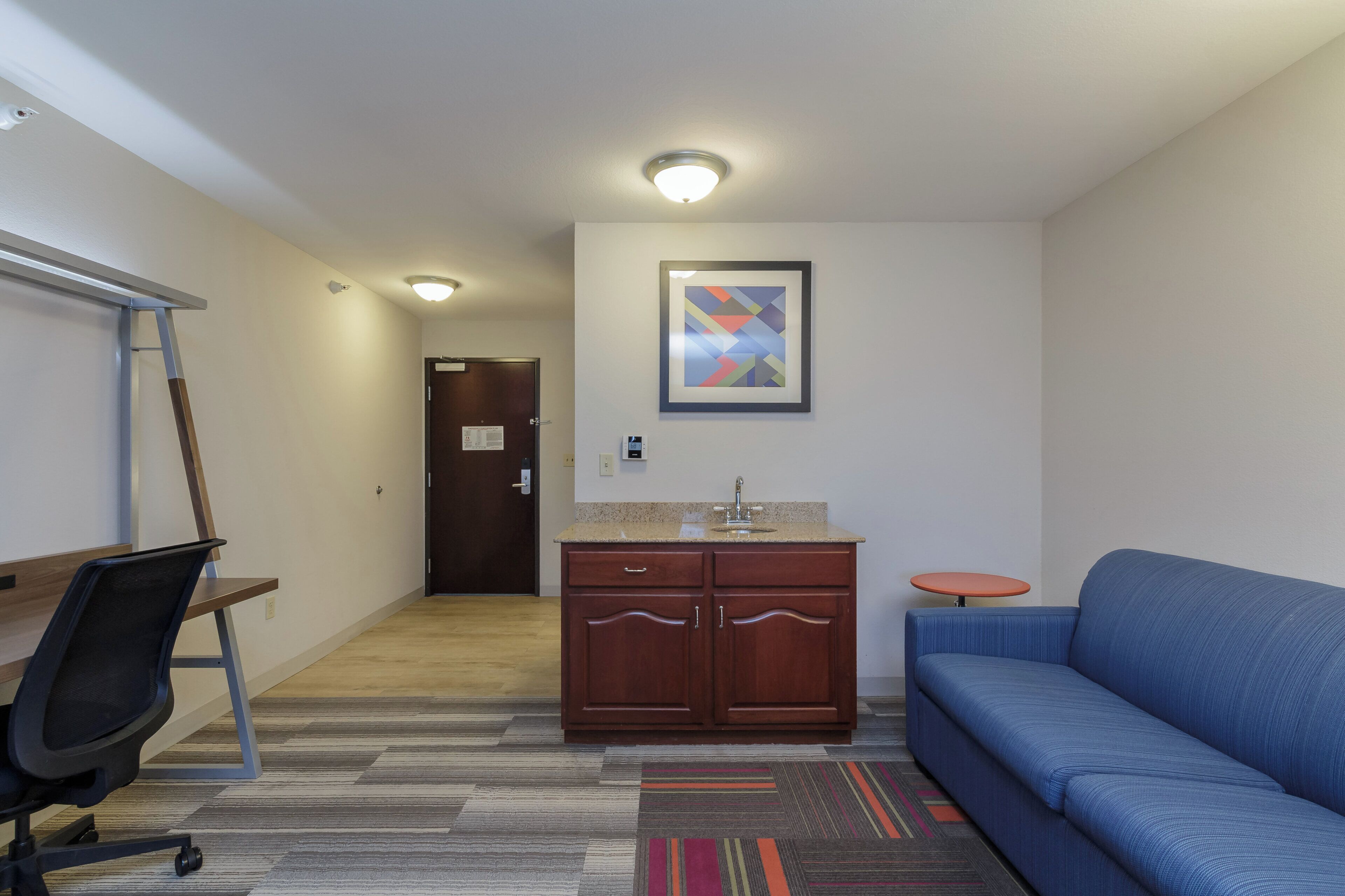 A hotel room with a kitchenette, a blue sofa, a small table, a chair, and a carpeted floor.