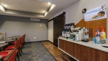 Habitación de hotel con comedor que incluye sillas y mesas rojas, una kitchenette con máquinas de café, y un letrero que dice "Smart Roast".