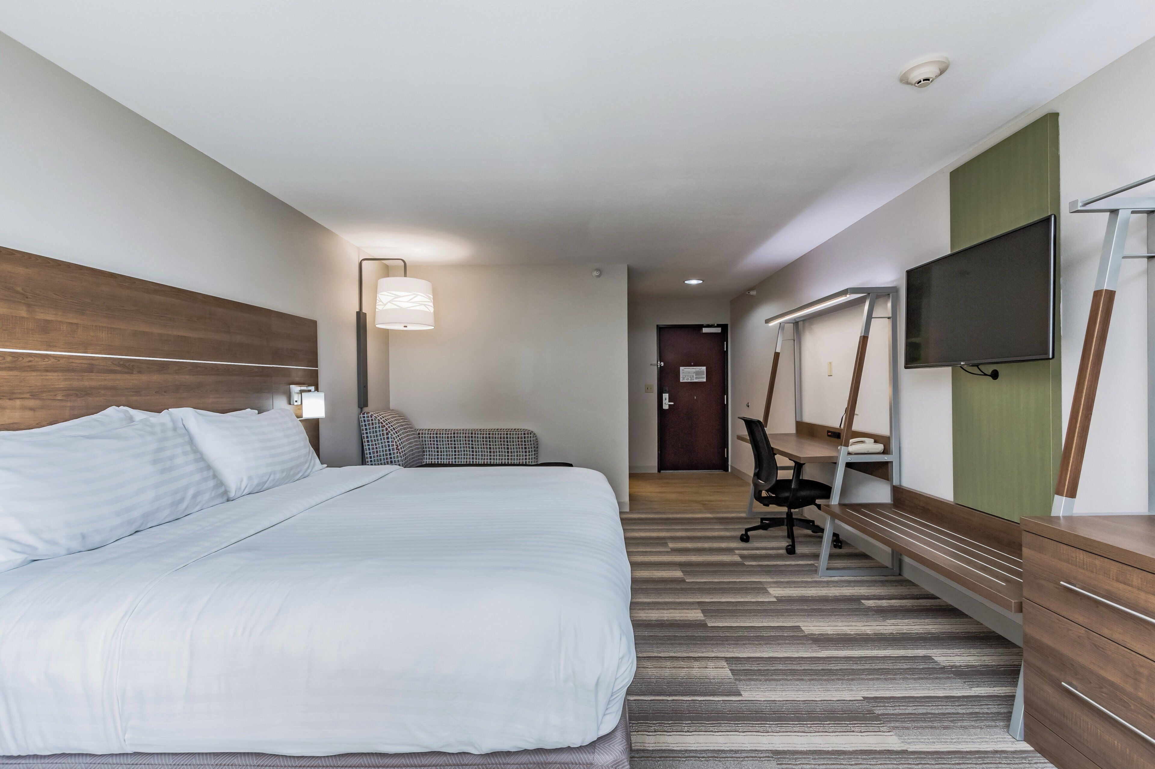 A hotel room with a large bed, a desk with a chair, a flat-screen TV, and a lamp.