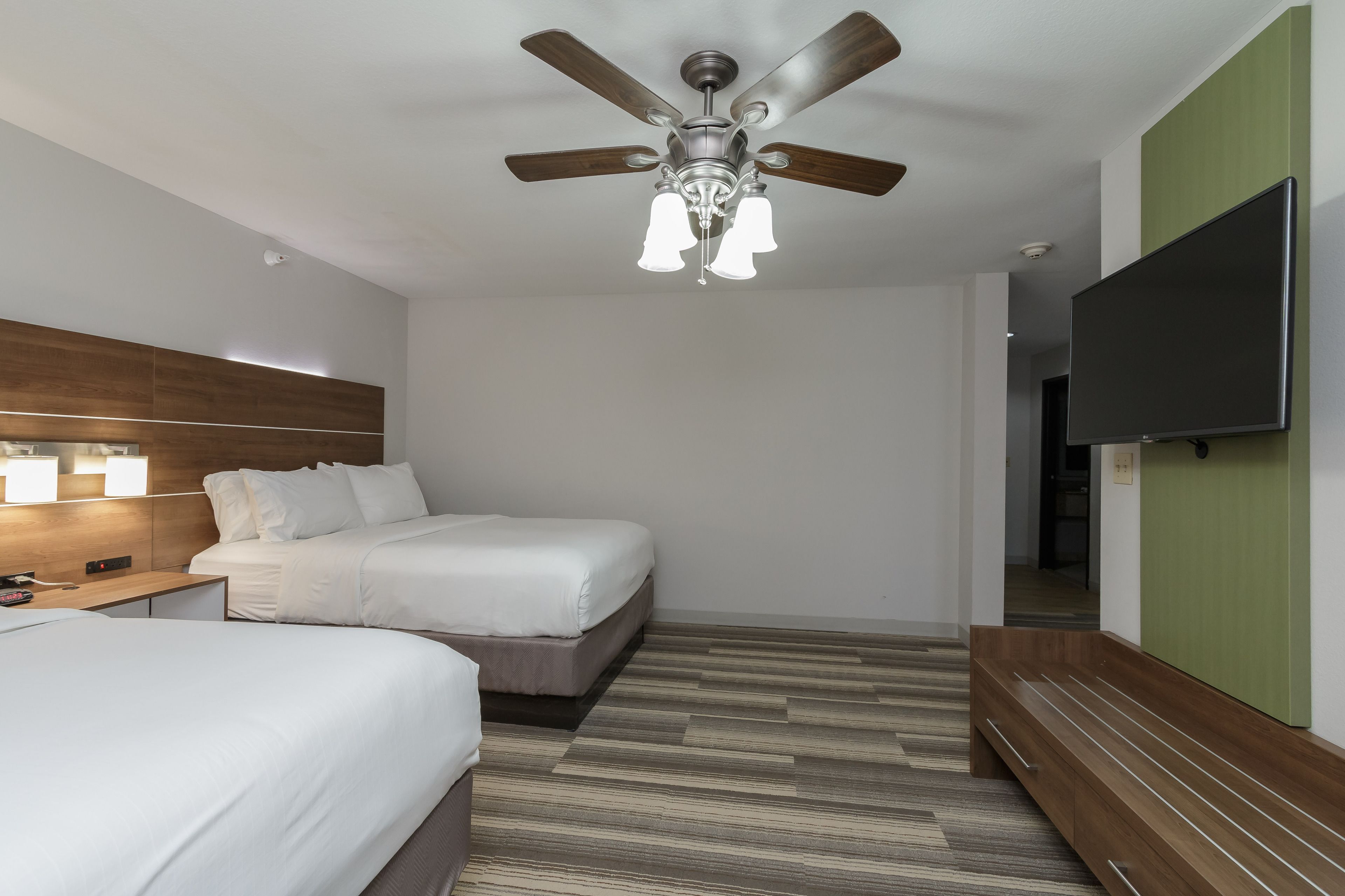 A hotel room with two beds, a ceiling fan, a flat-screen TV, and a wooden bench.