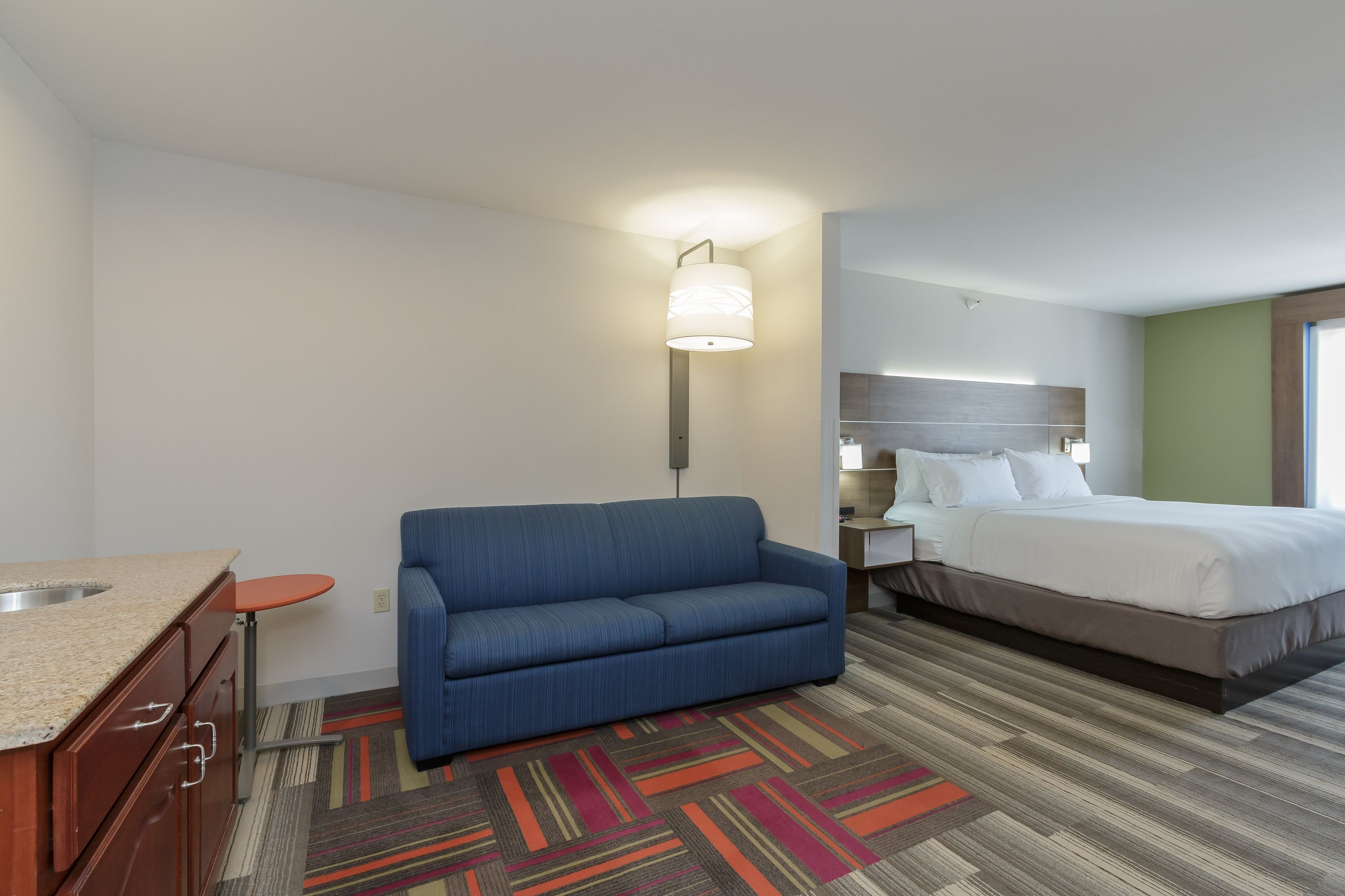 A hotel room with a kitchenette, a blue sofa, a bed with white linens, and a patterned rug.