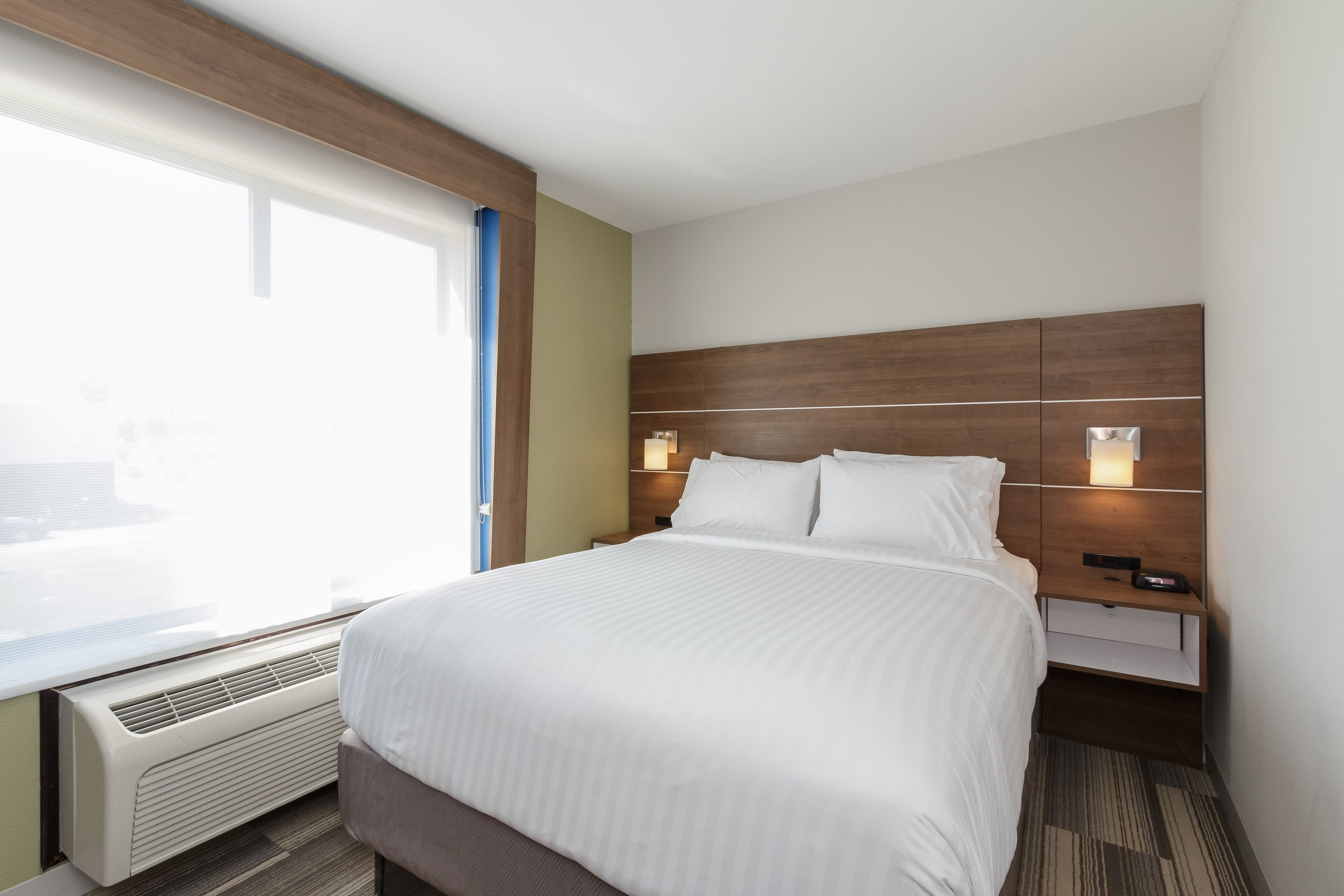 Une chambre d’hôtel avec un grand lit, une table de chevet avec une lampe, une fenêtre avec des stores et un radiateur fixé au mur.