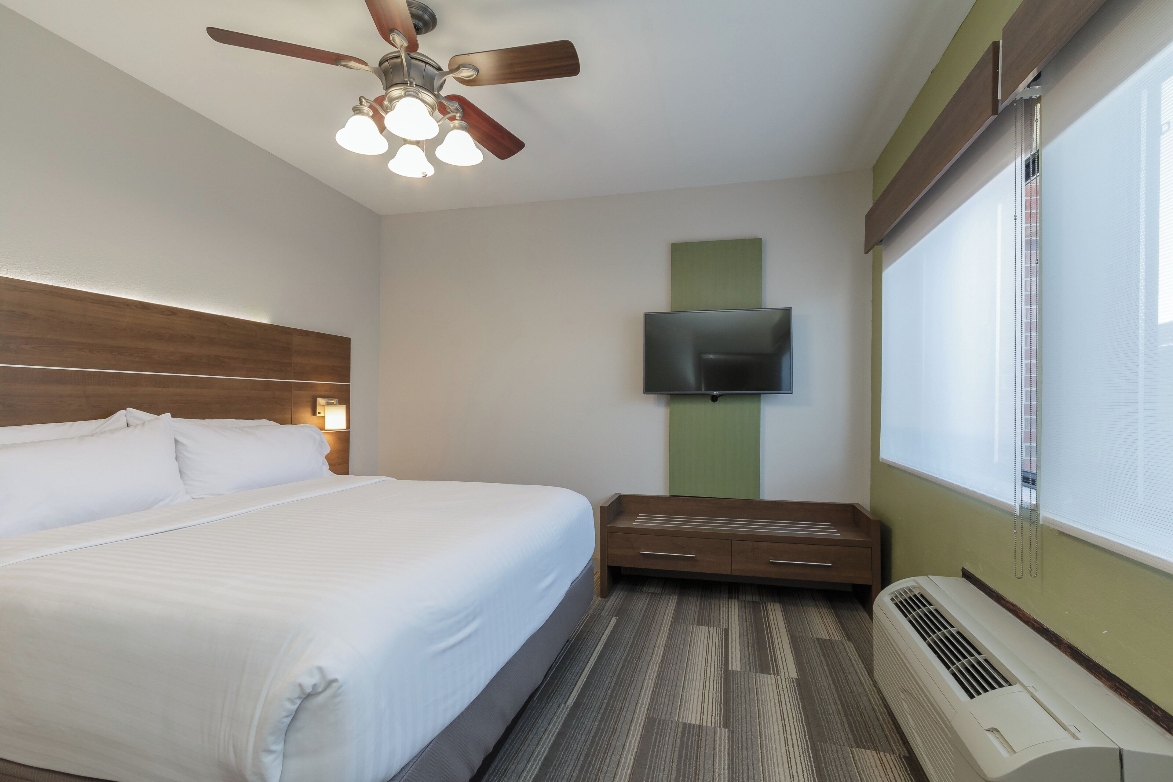 A hotel room with a bed, a flat-screen TV mounted on the wall, a ceiling fan, and an air conditioning unit.