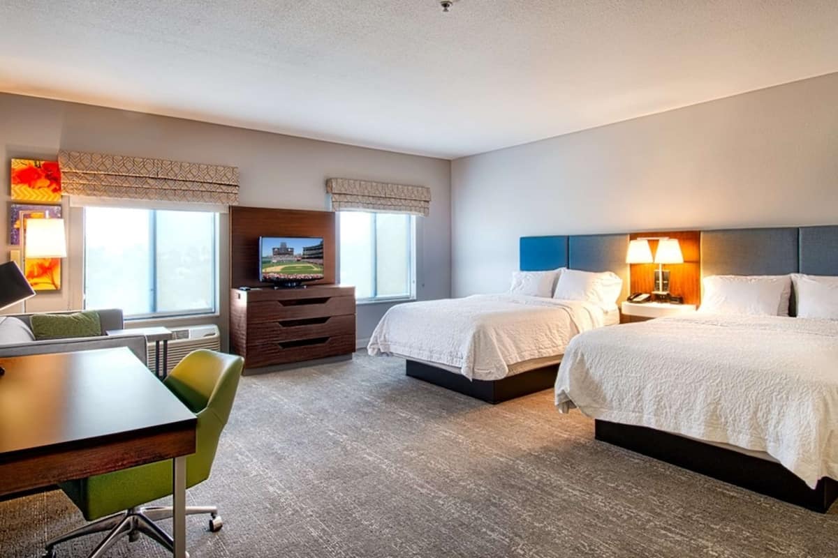 A hotel room with two beds, a desk, a chair, a TV, and a lamp.