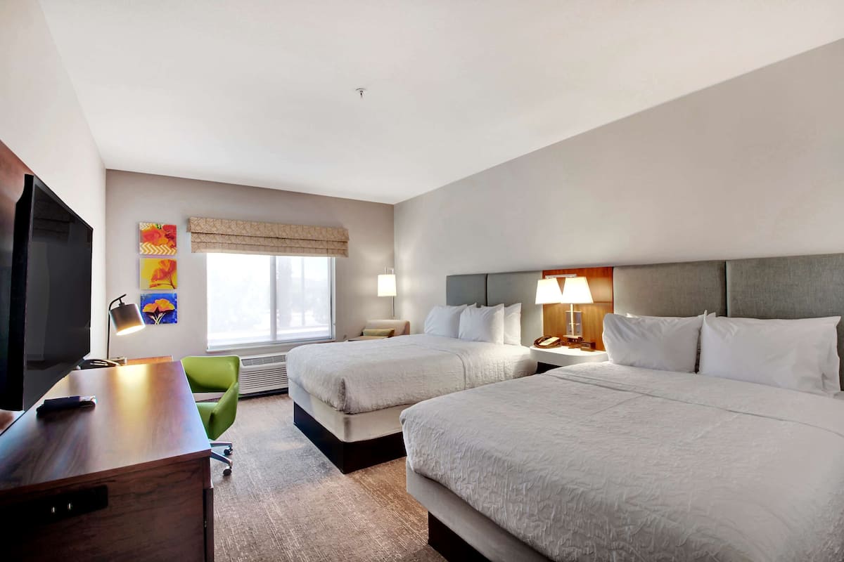 A hotel room with two beds, a desk, a TV, and a window with blinds.
