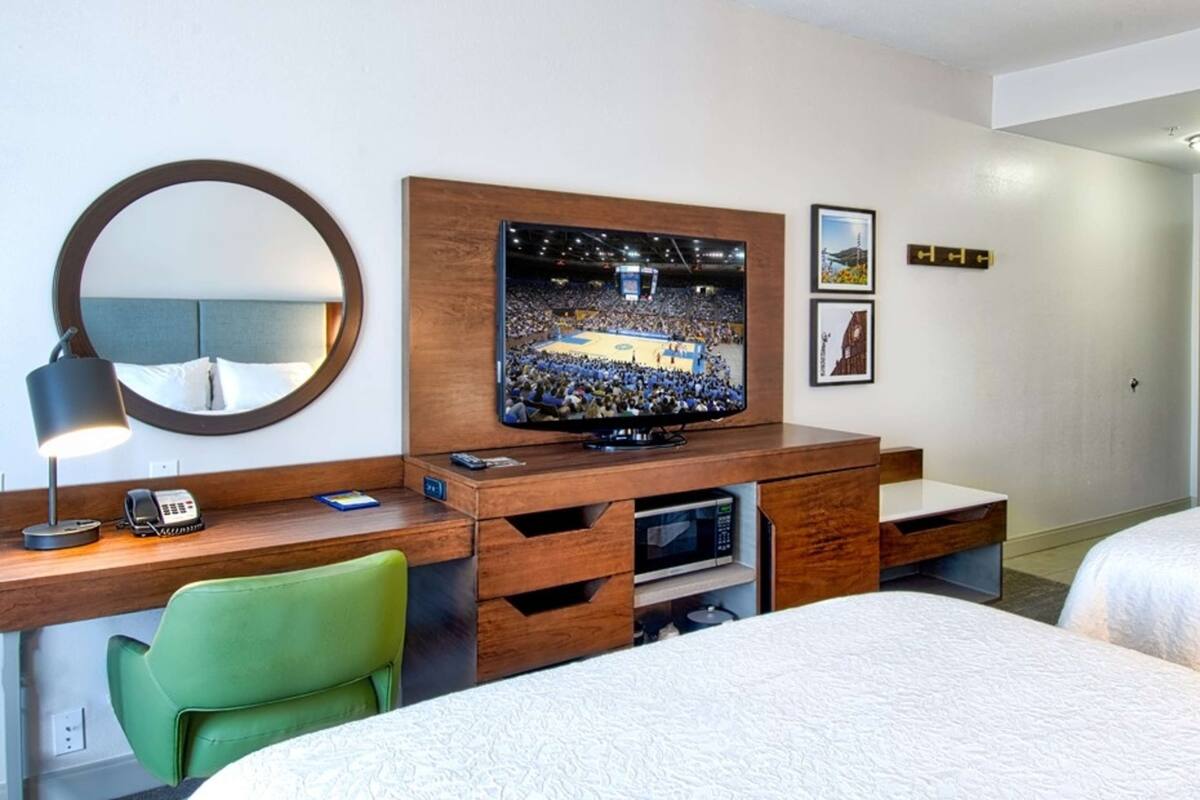 A hotel room with a bed, a desk with a computer, a TV, a mirror, and a green chair.