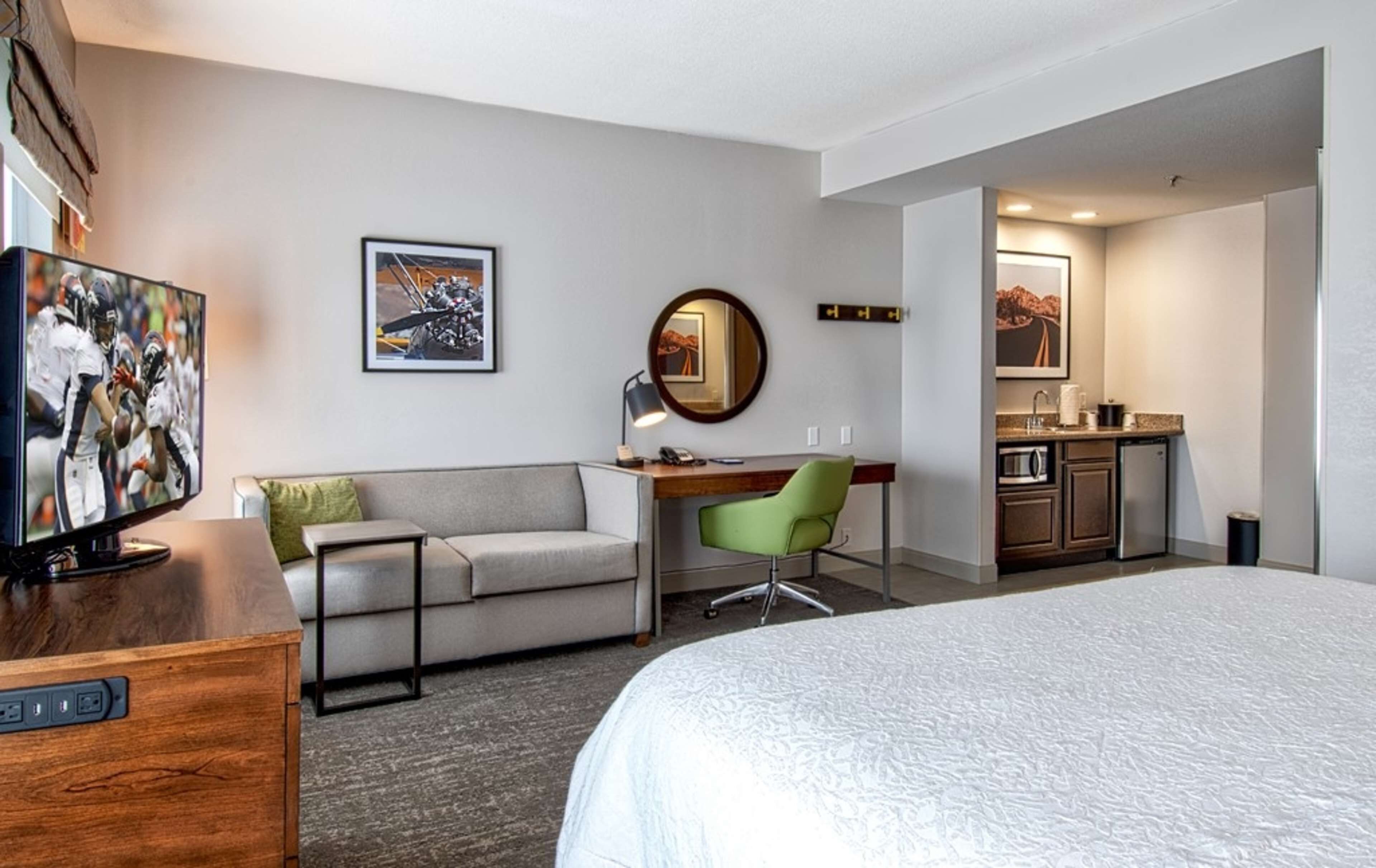 A hotel room with a bed, a desk with a chair, a sofa, a television, a mirror, and a kitchenette.