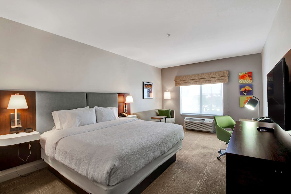 A hotel room with a large bed, bedside lamps, a desk with a TV, a chair, and a window with blinds.