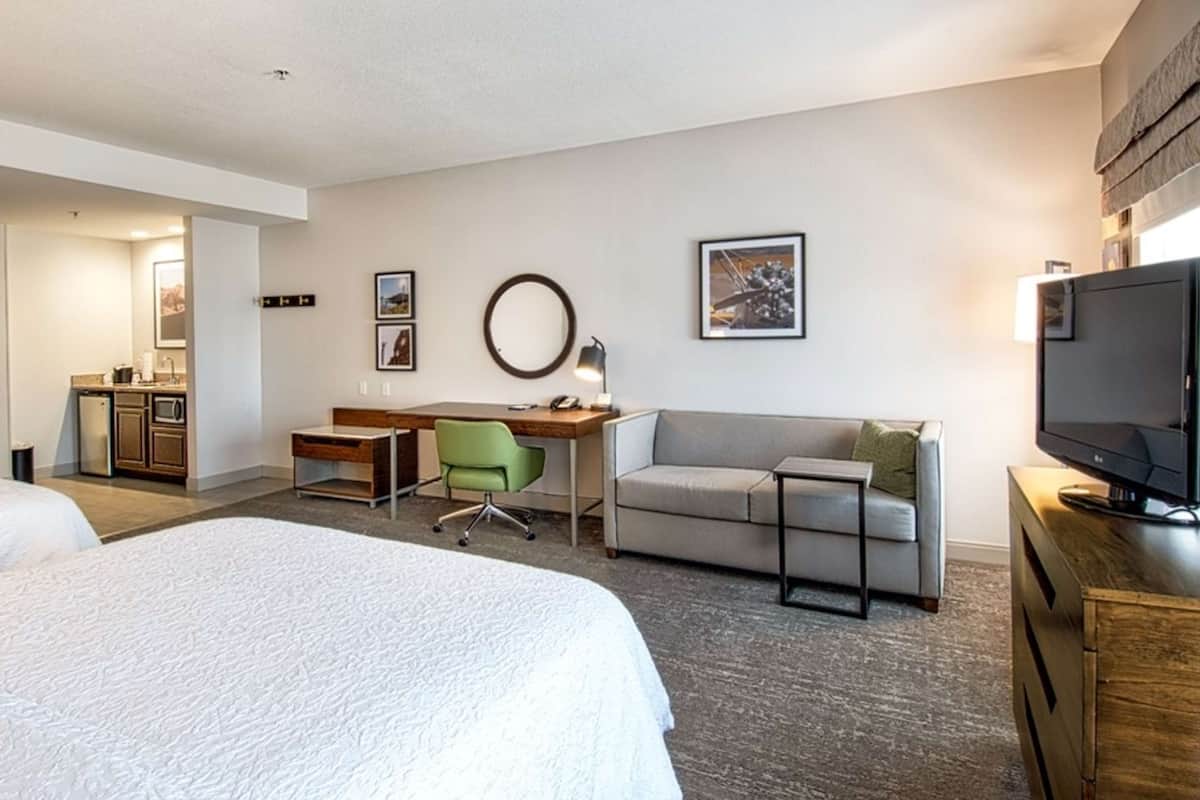A hotel room with two beds, a desk, a chair, a sofa, a TV, and a kitchenette.