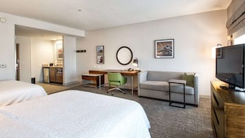 A hotel room with two beds, a desk, a chair, a sofa, a TV, and a kitchenette.