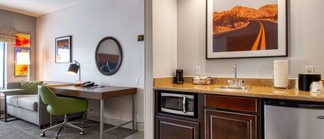 Une chambre d’hôtel compacte comprenant un coin cuisine, un bureau, un fauteuil de bureau vert, un miroir rond et un tableau représentant une route du désert, accroché au mur.