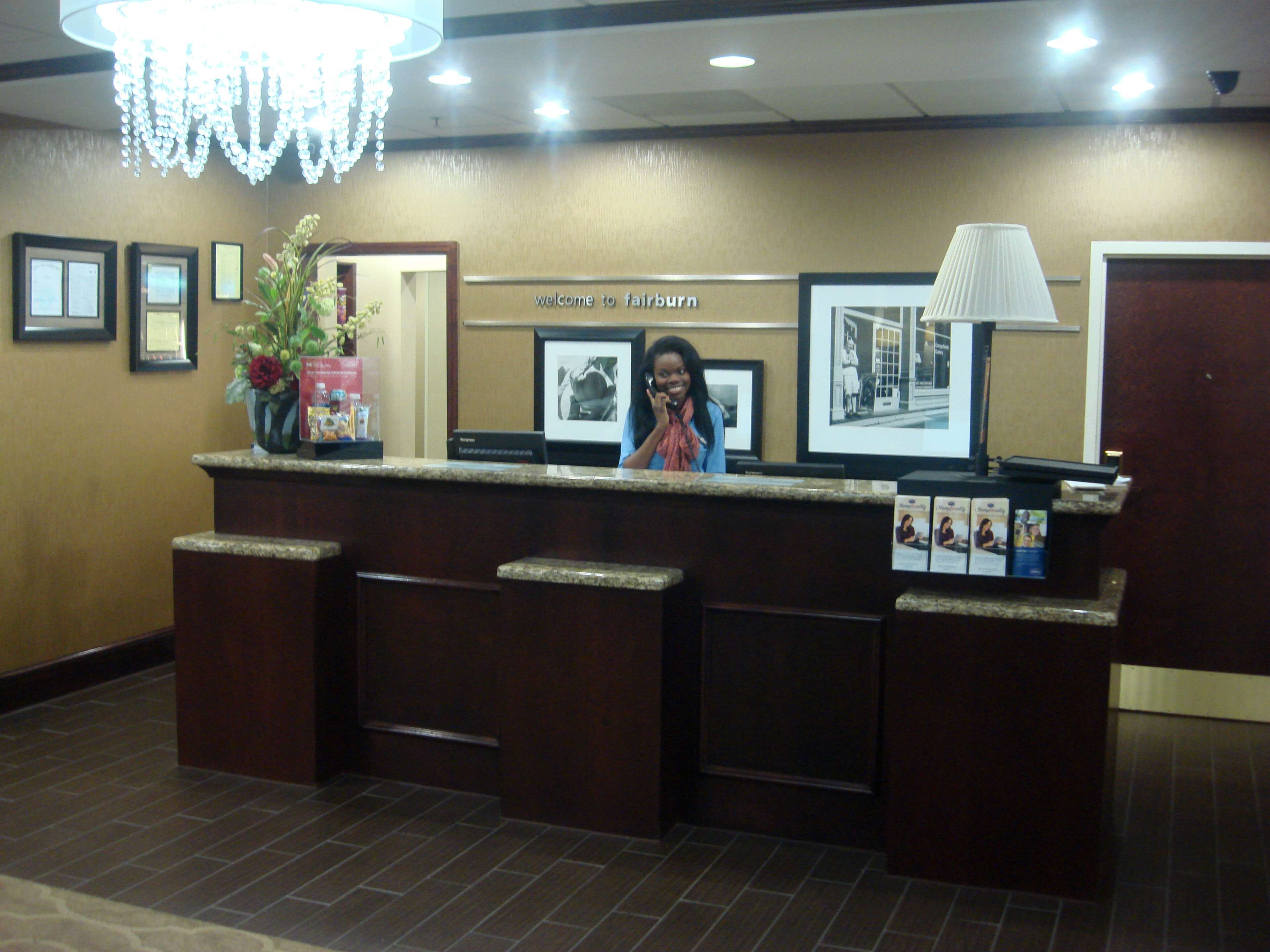 A reception desk with a 'Welcome to Fairburn' sign, framed pictures on the wall, a potted plant, and a lamp.