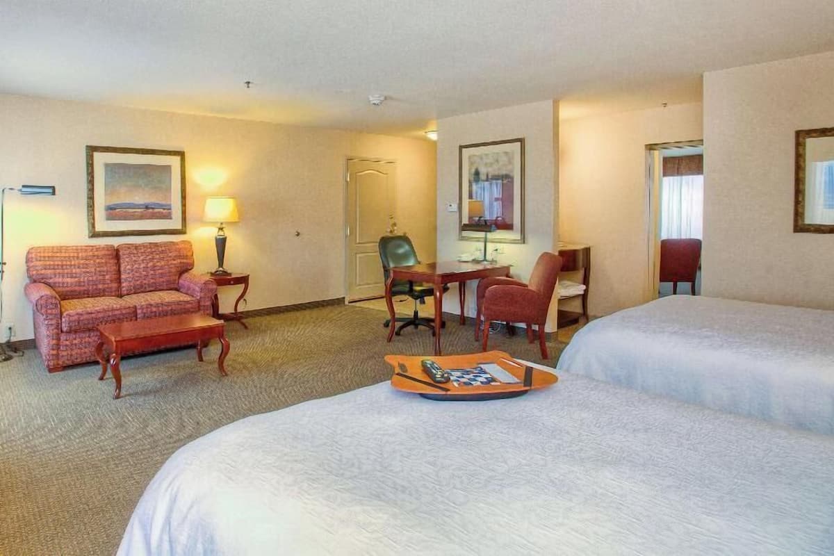 Quarto de hotel com duas camas, uma escrivaninha, uma cadeira, um sofá e um abajur.