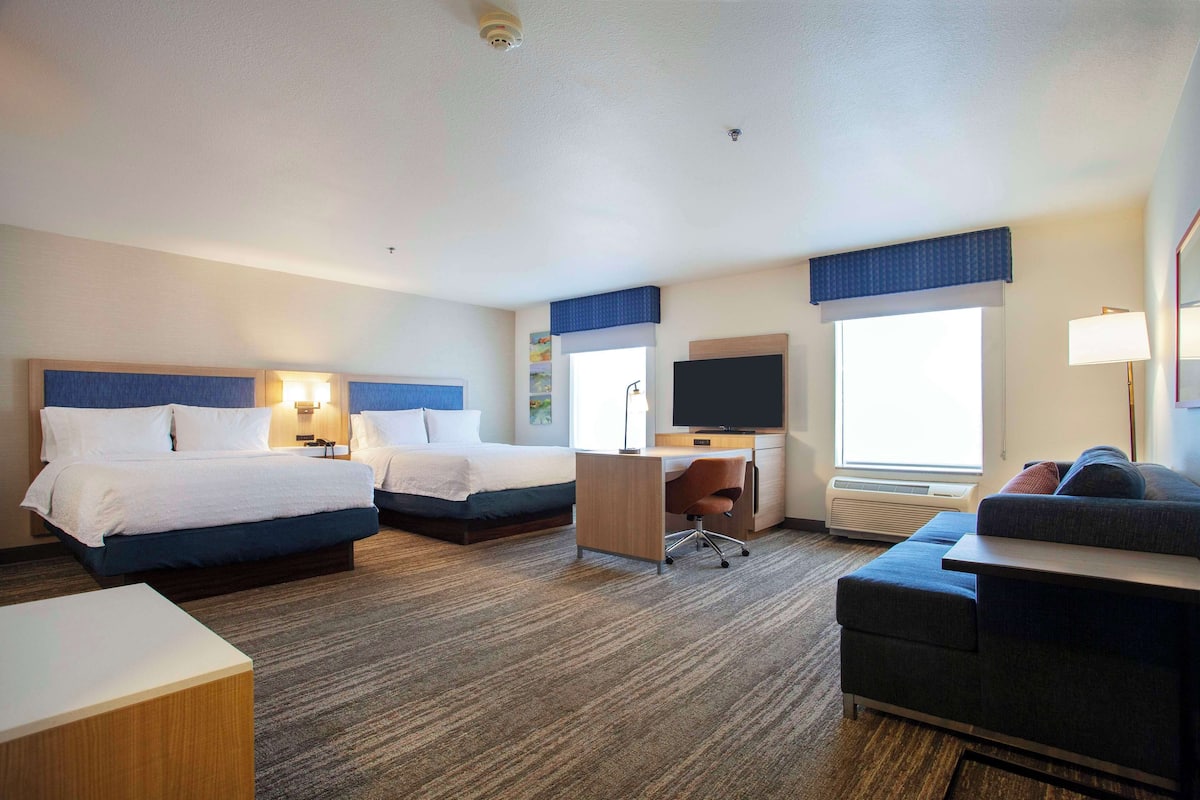 Quarto de hotel com duas camas, uma escrivaninha, uma televisão e um sofá.