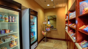 A hotel corridor with a refrigerated display case on the left, a snack vending machine in the center, and shelves stocked with snacks on the right. - Hampton Inn & Suites Mountain Home (Mountain Home)