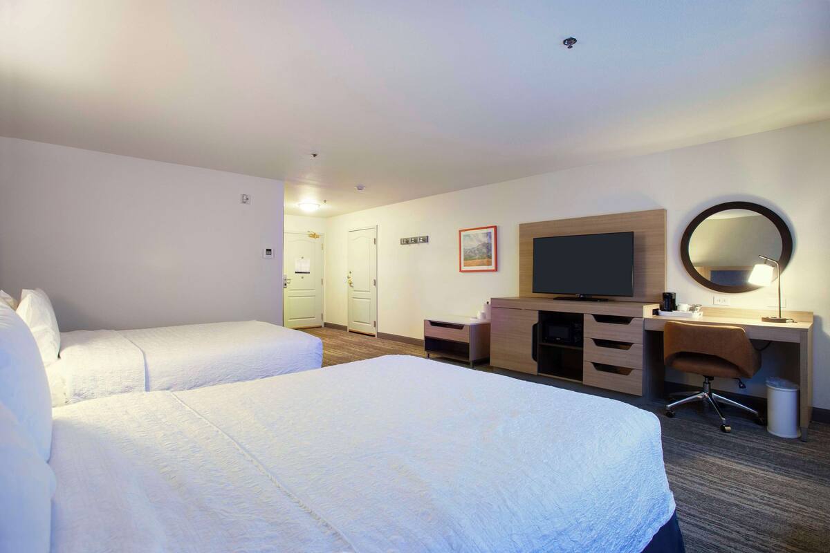 Quarto de hotel com duas camas, uma escrivaninha, uma cadeira, uma televisão e um espelho.