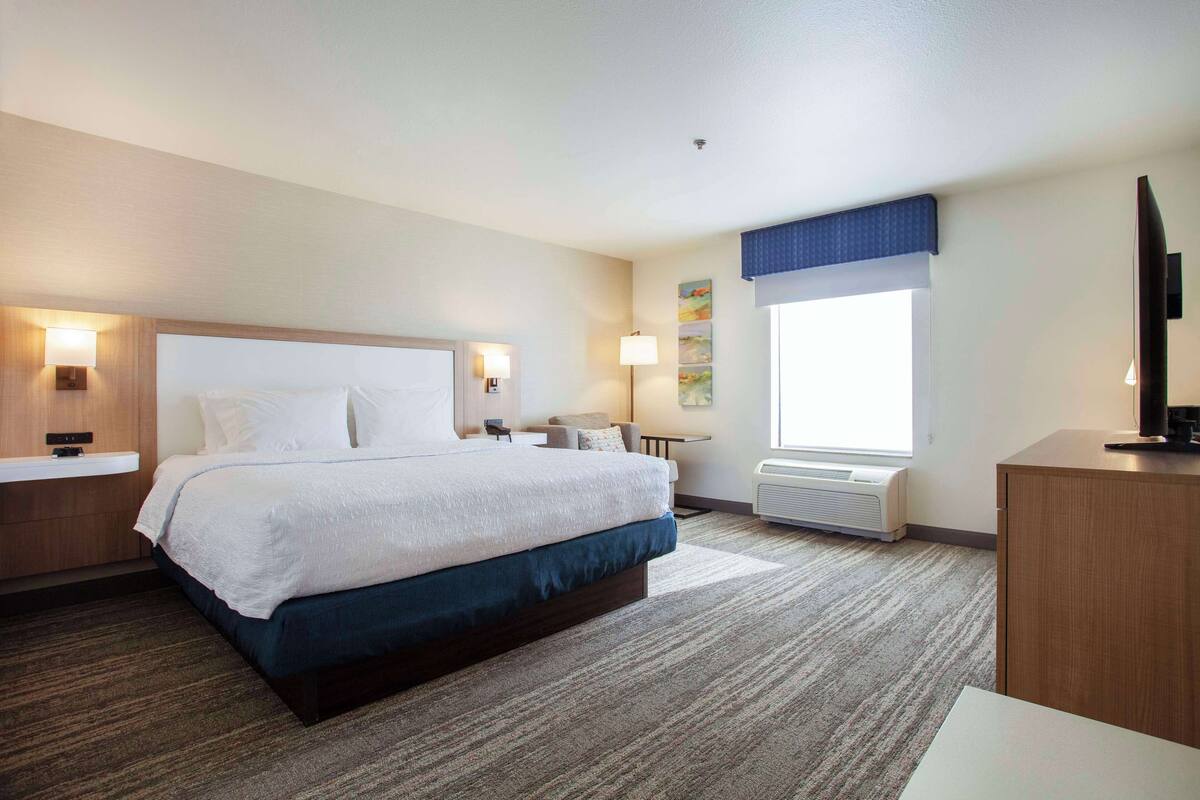 Quarto de hotel com uma cama grande, luminárias de cabeceira, uma escrivaninha e uma janela com persianas.