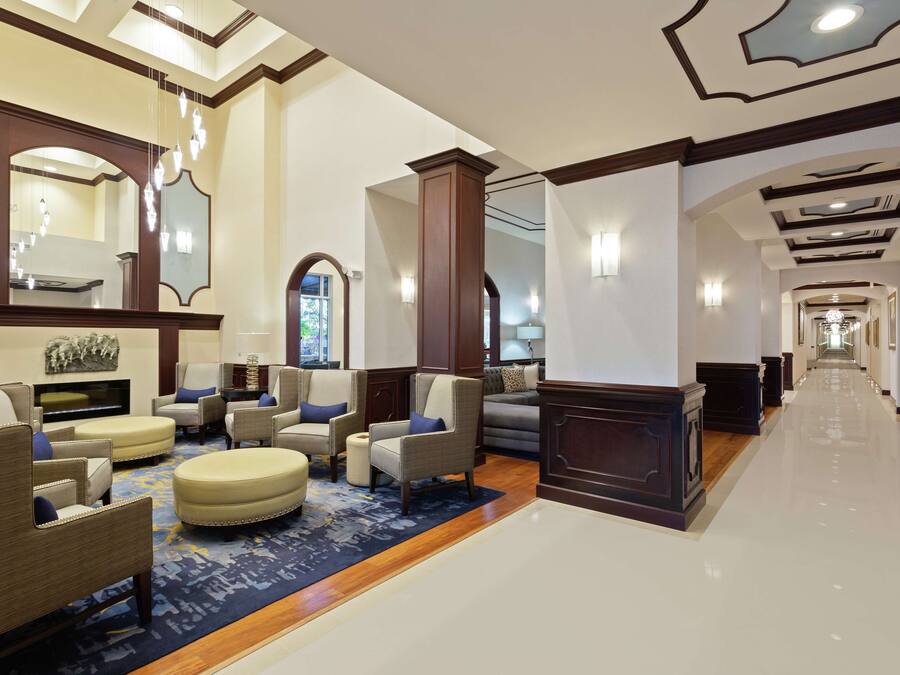 A spacious lobby with a high ceiling, a central seating area with sofas and armchairs, a reception desk, and a long corridor leading to other areas.