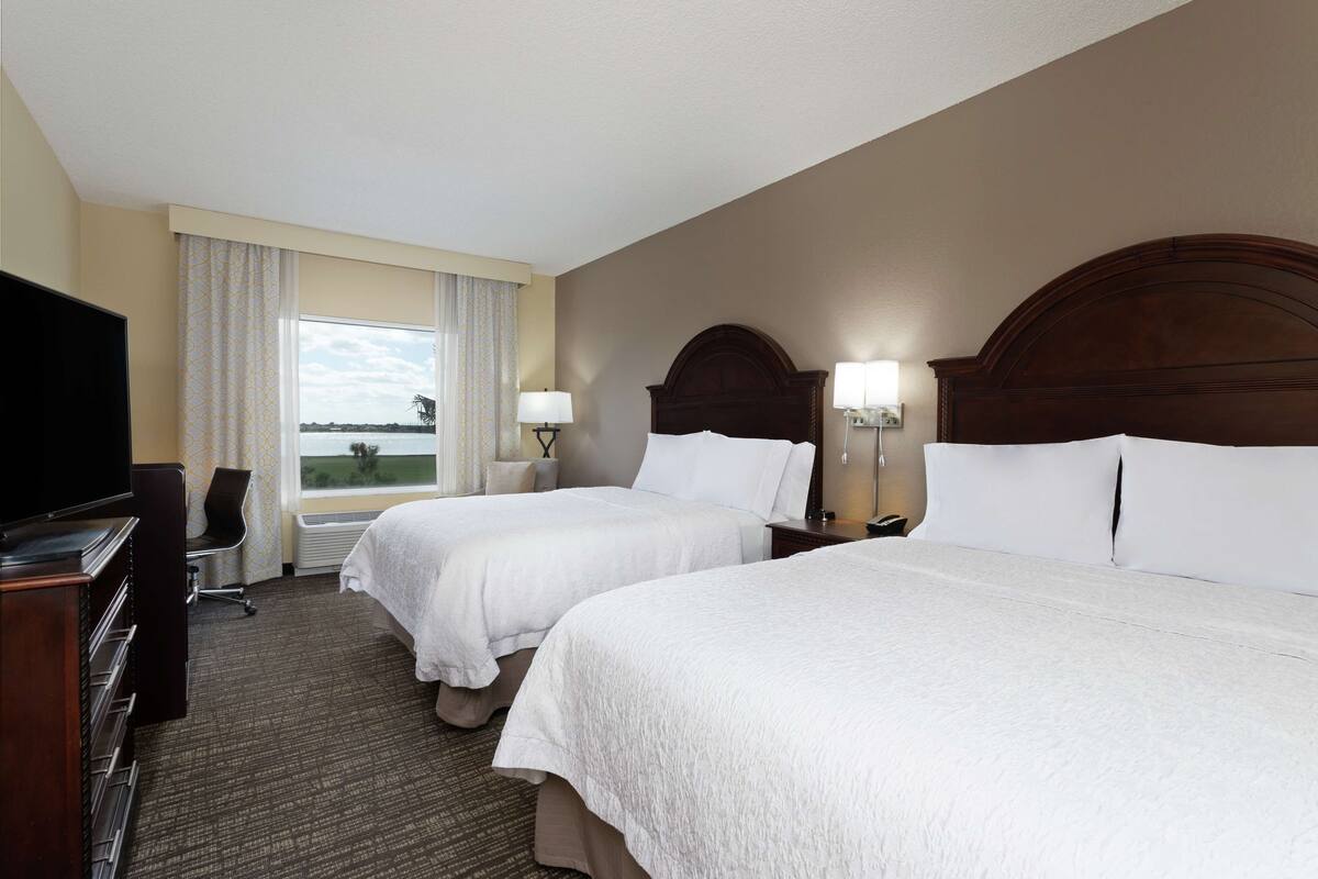 A hotel room with two beds, a TV, a desk, and a window with curtains.