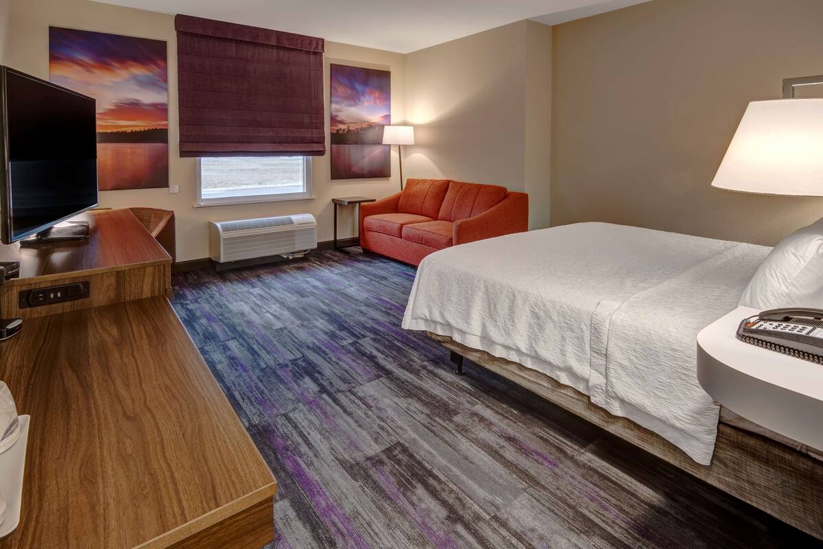 A hotel room with a bed, a TV, a desk, a chair, and a window with a view of a sunset.