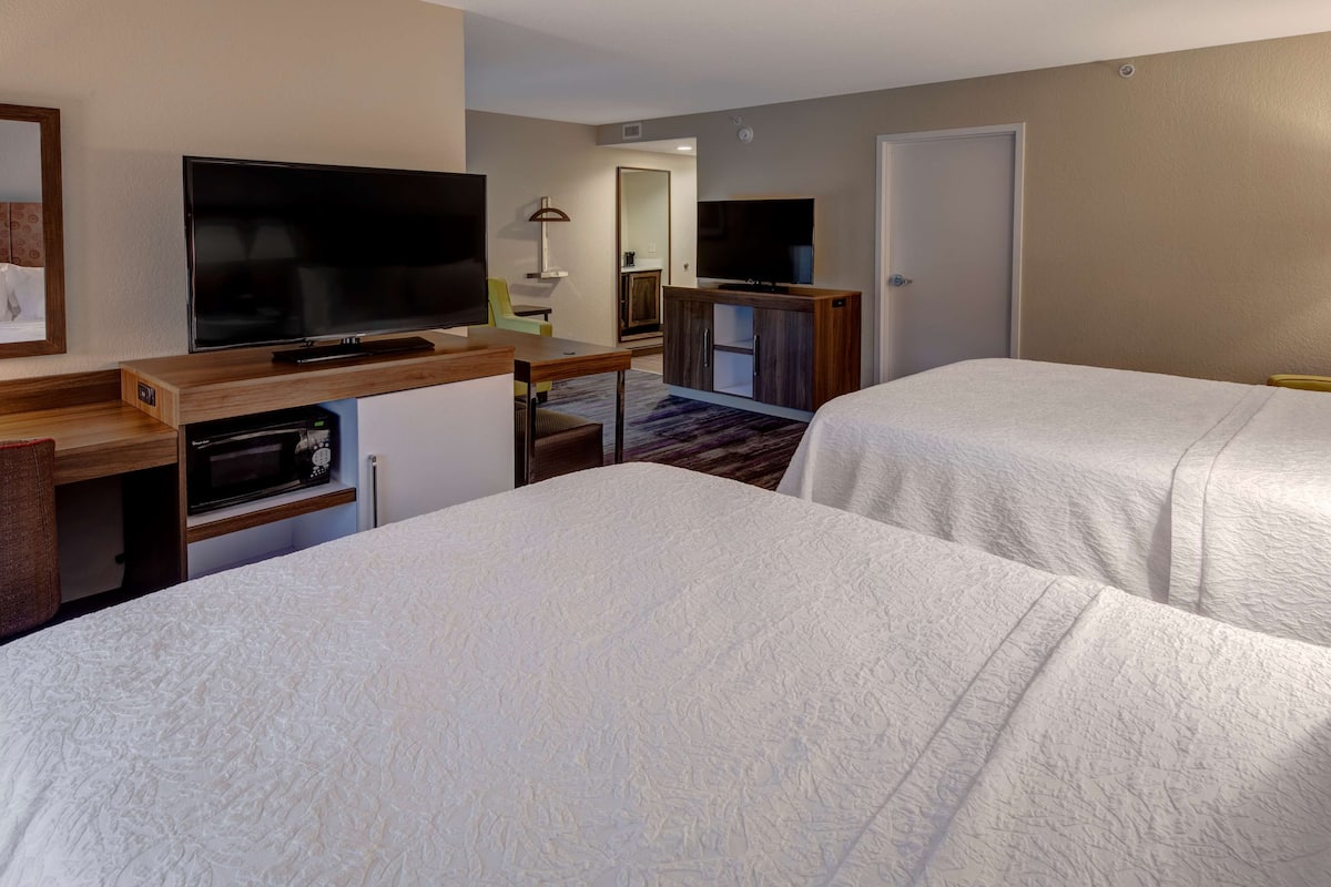 A hotel room with two beds, a TV, a desk with a computer, and a small kitchenette.