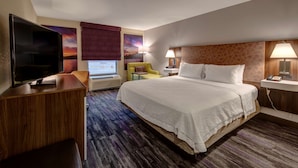 Une chambre d’hôtel avec un grand lit, un téléviseur à écran plat, un bureau avec une chaise, une petite table et une fenêtre donnant sur un coucher de soleil.