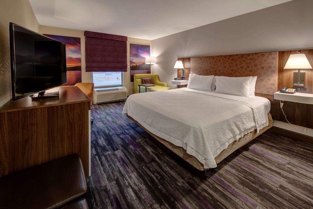 A hotel room with a large bed, a flat-screen TV, a desk with a chair, a small table, and a window with a view of a sunset.