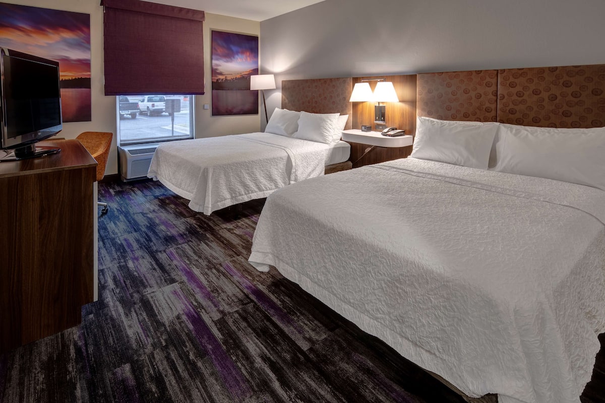 A hotel room with two beds, a TV, a desk, and a window with a view of parked cars.