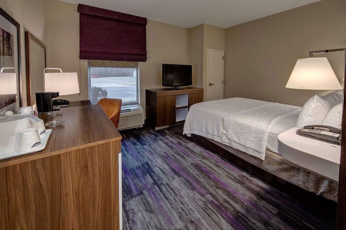 A hotel room with a bed, a desk with a chair, a TV, a bathroom with a sink and mirror, and a window with a view of a parking lot.