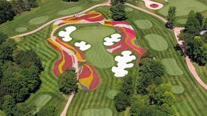 An aerial view of a golf course with multiple greens, sand bunkers, and a central area with colorful flower arrangements.