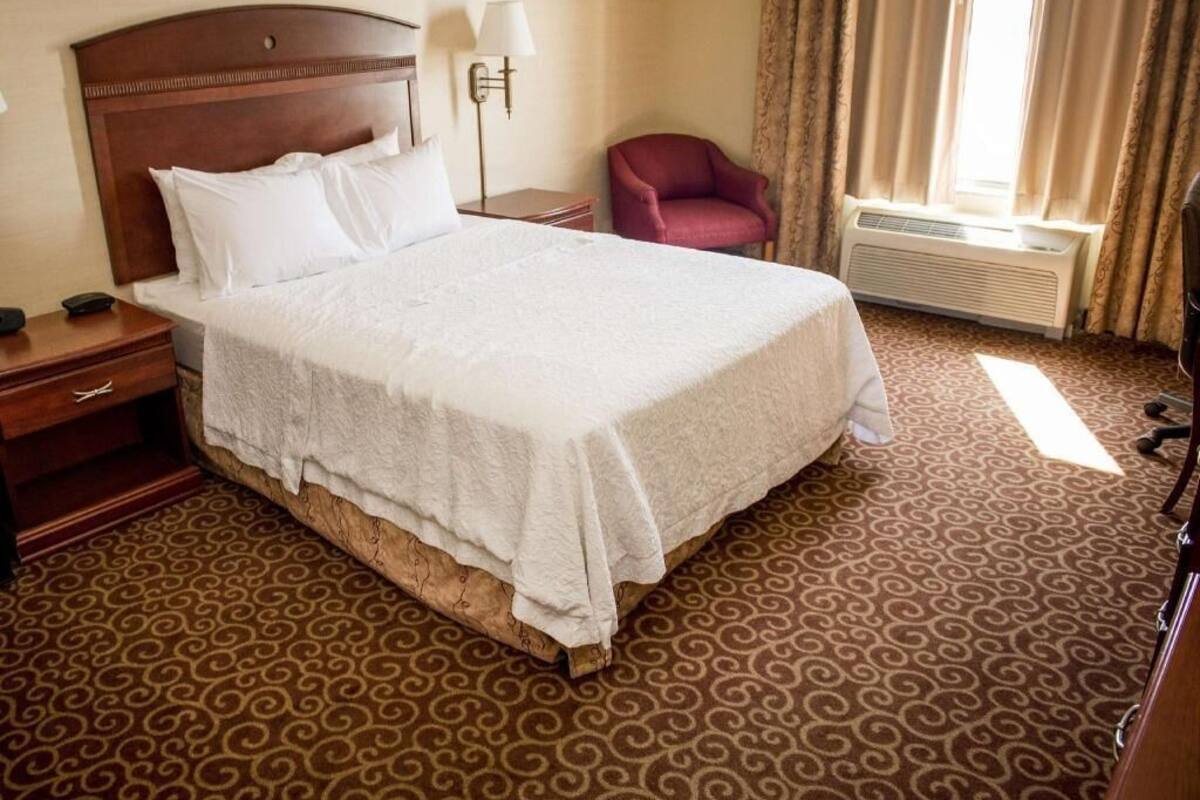 Ein Hotelzimmer mit einem großen Bett, einem roten Sessel, einem Nachttisch mit Lampe und einem fenster mit Vorhängen.