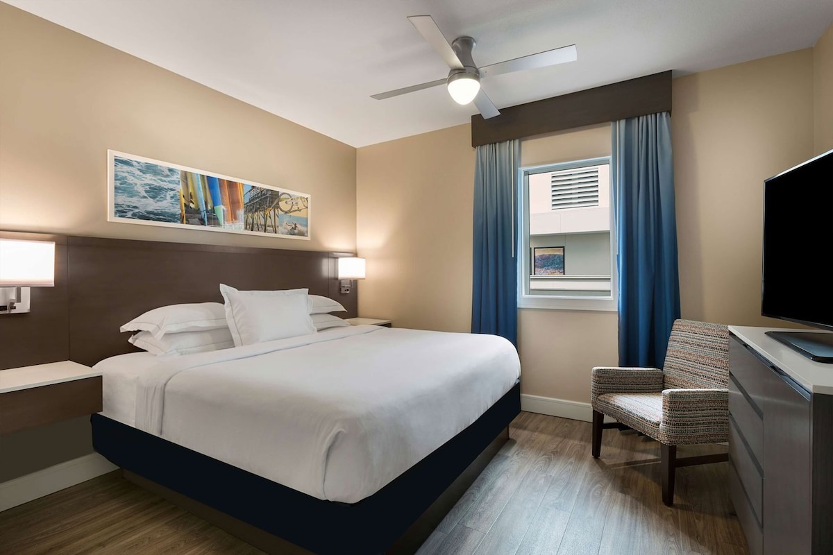 Ein Hotelzimmer mit Bett, Schreibtisch mit Flachbildfernseher, Stuhl, Deckenventilator, Fenster mit Vorhängen und einem Bild an der Wand.