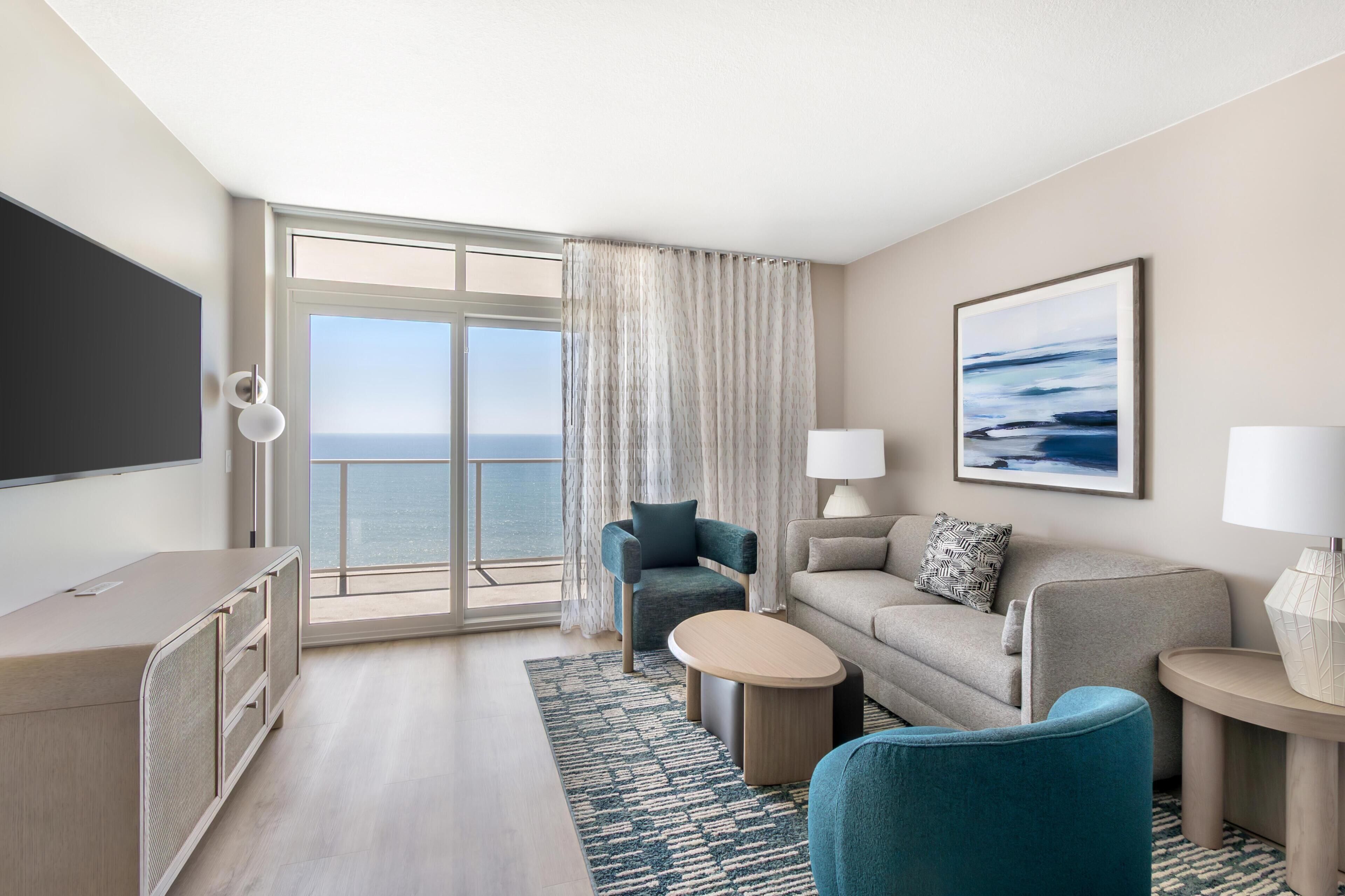 Une chambre d’hôtel moderne avec une grande fenêtre, un canapé, un fauteuil, une petite table et un téléviseur.