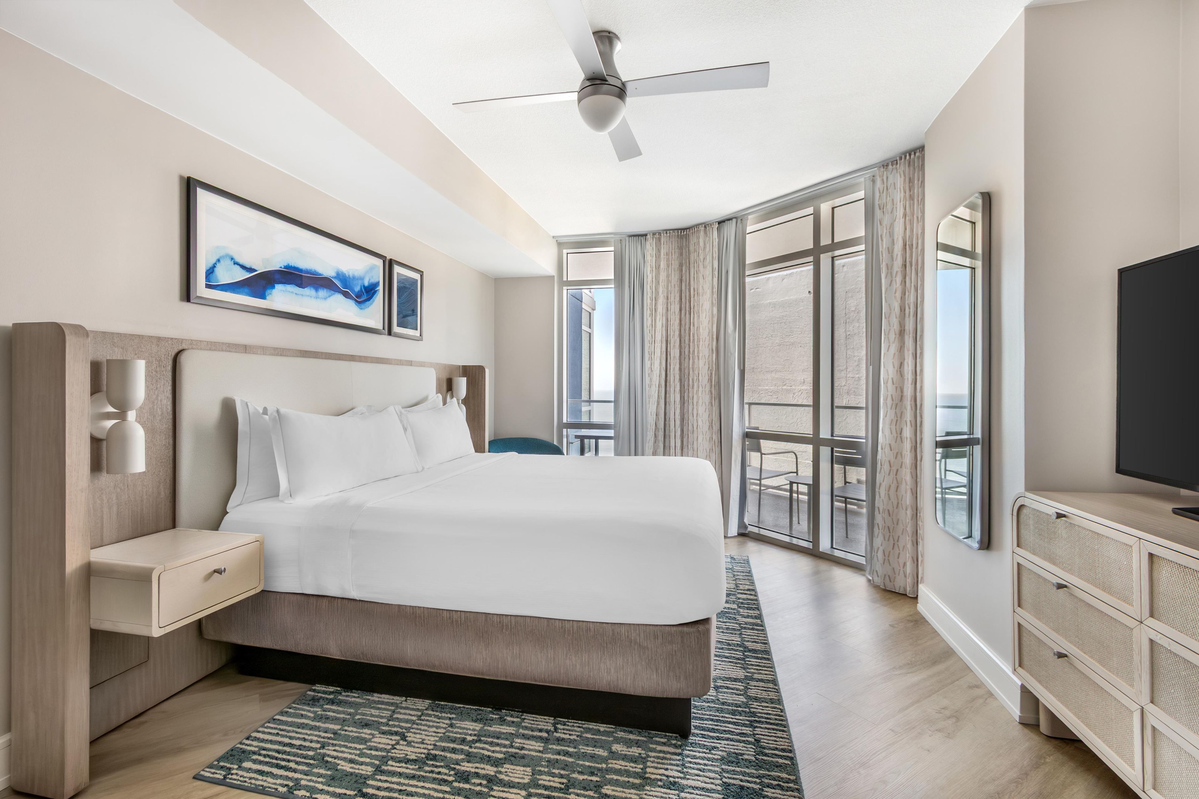 A hotel room with a bed, a TV, a ceiling fan, a nightstand, a desk, and a large window.