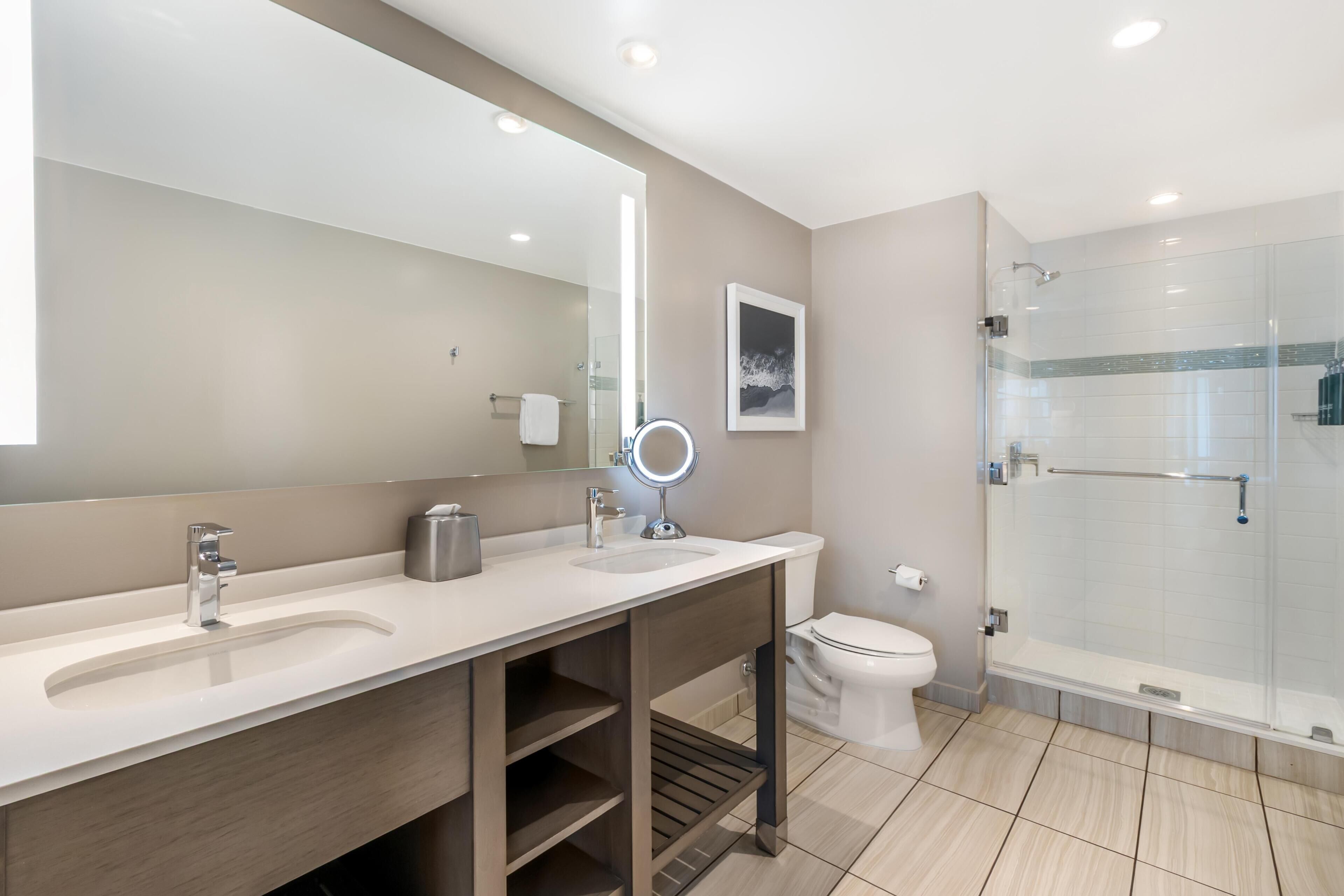A modern bathroom with a large mirror, two sinks, a toilet, and a glass-enclosed shower.