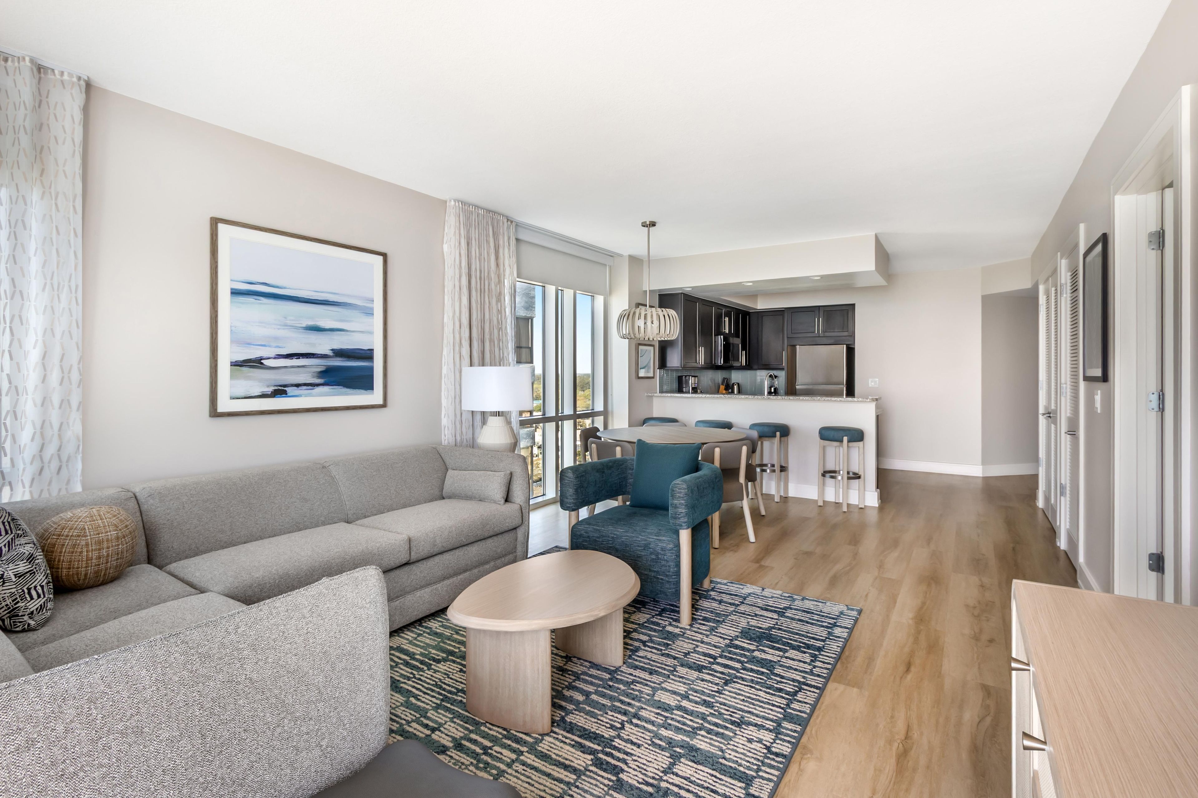 A modern living room with a sofa, coffee table, and chairs, a dining area with a table and chairs, and a kitchen with a refrigerator and stove.