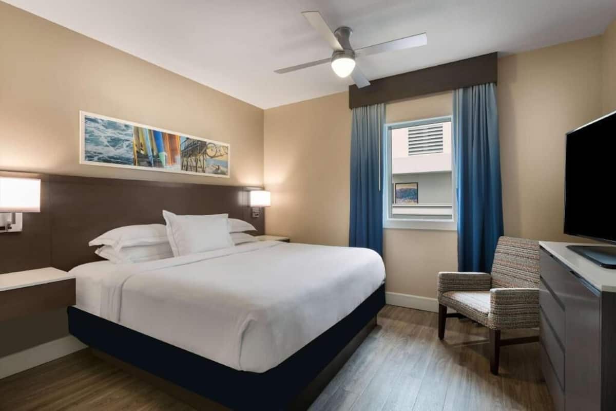 Ett hotellrum med en säng, ett skrivbord med en stol, en takfläkt och ett fönster med gardiner.