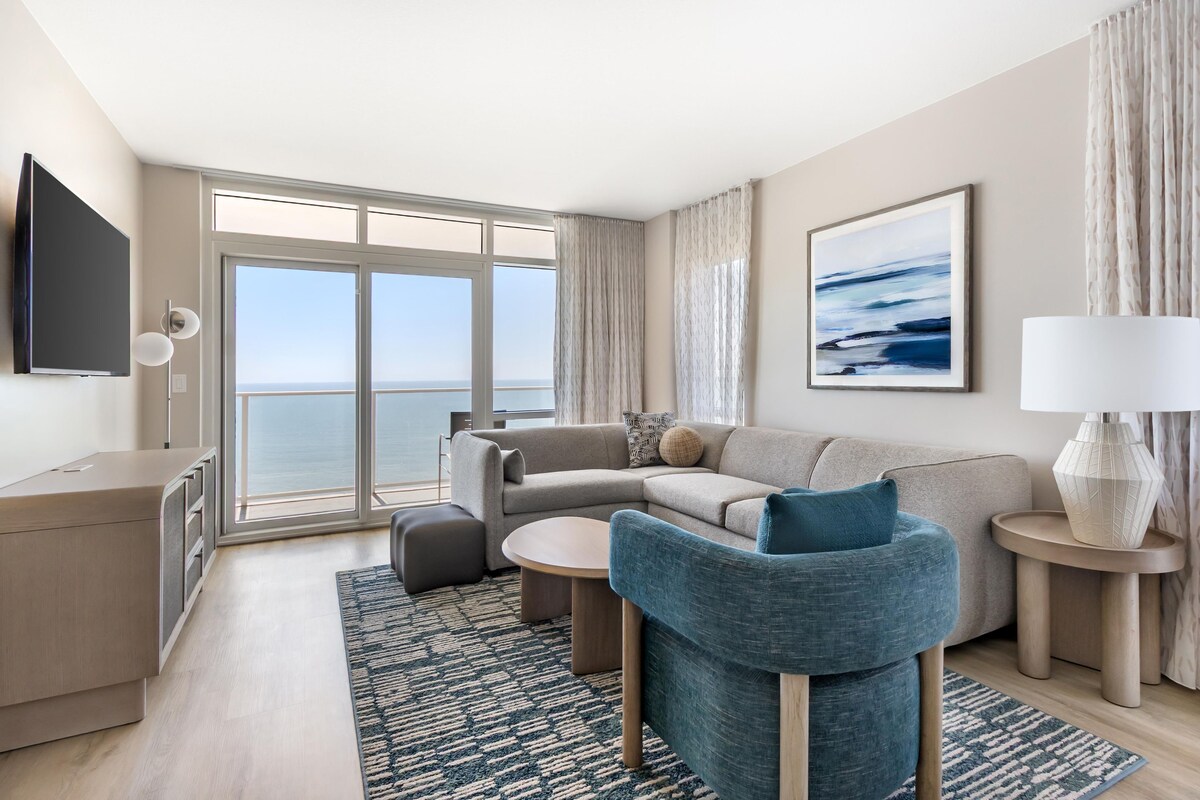 Una habitación de hotel moderna con un ventanal grande, un sofá, una silla, una mesita, una lámpara y un televisor de pantalla plana.