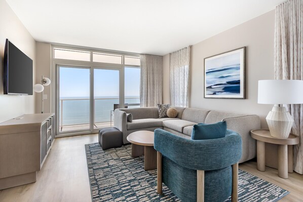 Una habitación de hotel moderna con un ventanal grande, un sofá, una silla, una mesita, una lámpara y un televisor de pantalla plana.