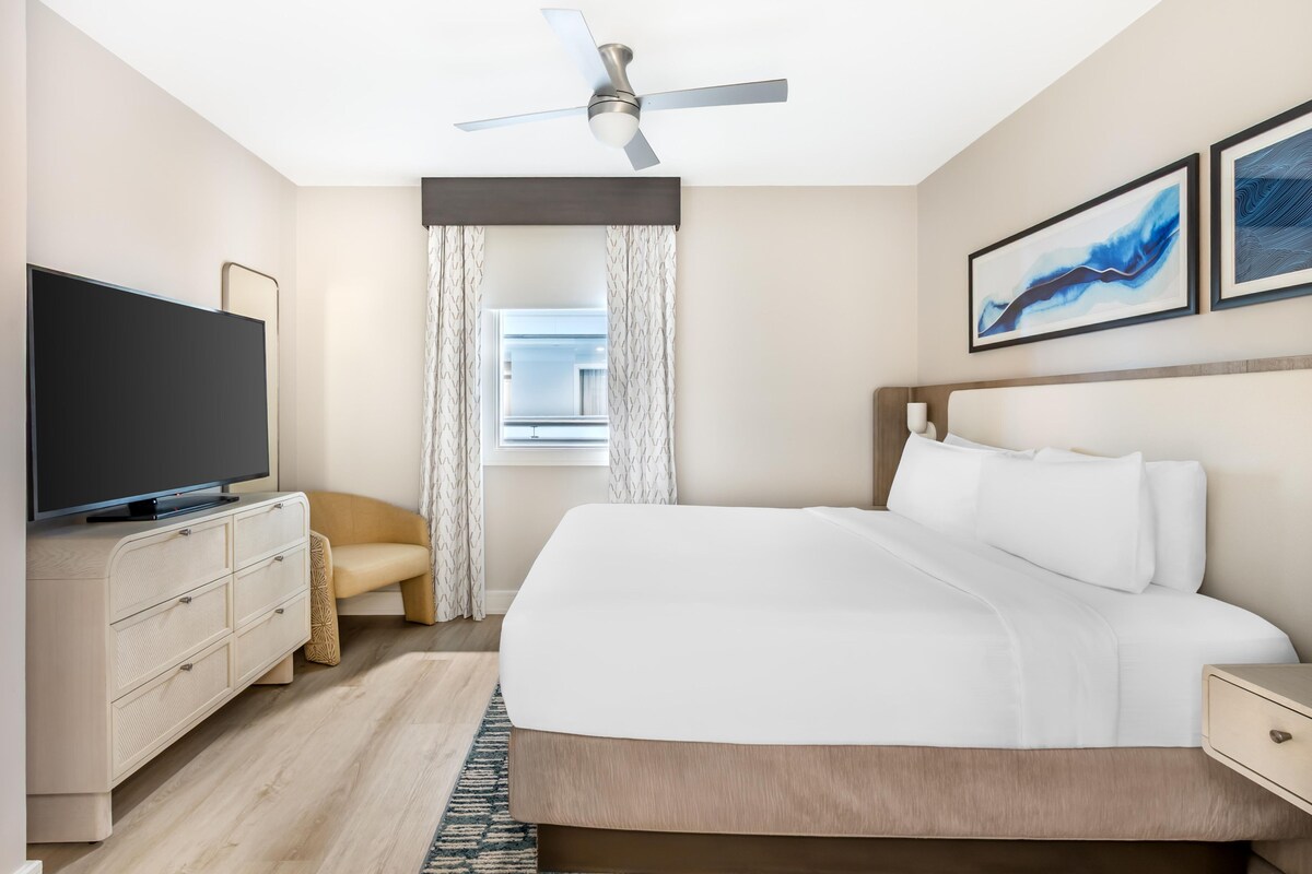 Una habitación de hotel con una cama, televisor, ventilador de techo, ventana con cortinas y una obra de arte en la pared.
